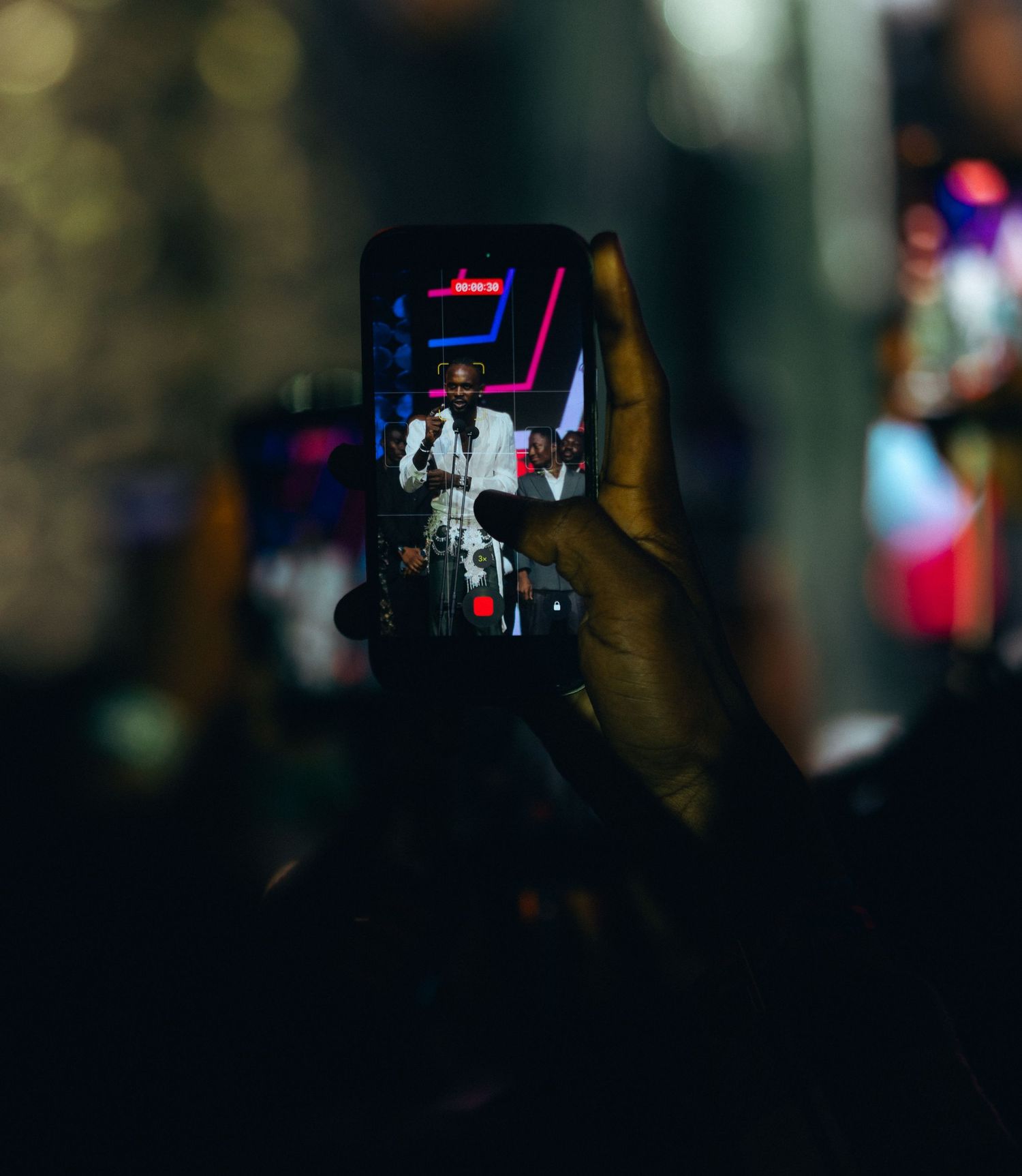 Concert fan records live performance on smartphone in dark venue with bright stage lights.
