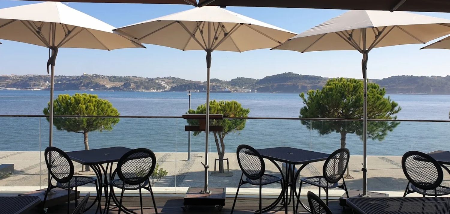 Beachfront restaurant terrace with white umbrellas and dining tables overlooking a calm ocean bay and distant mountains.