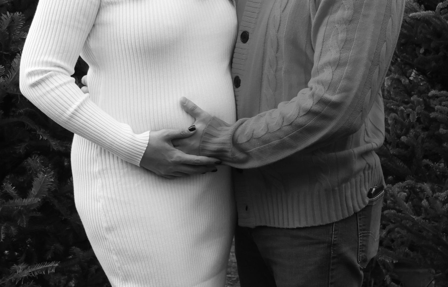 Black and white maternity photo of couple holding pregnant belly outdoors near evergreen trees.