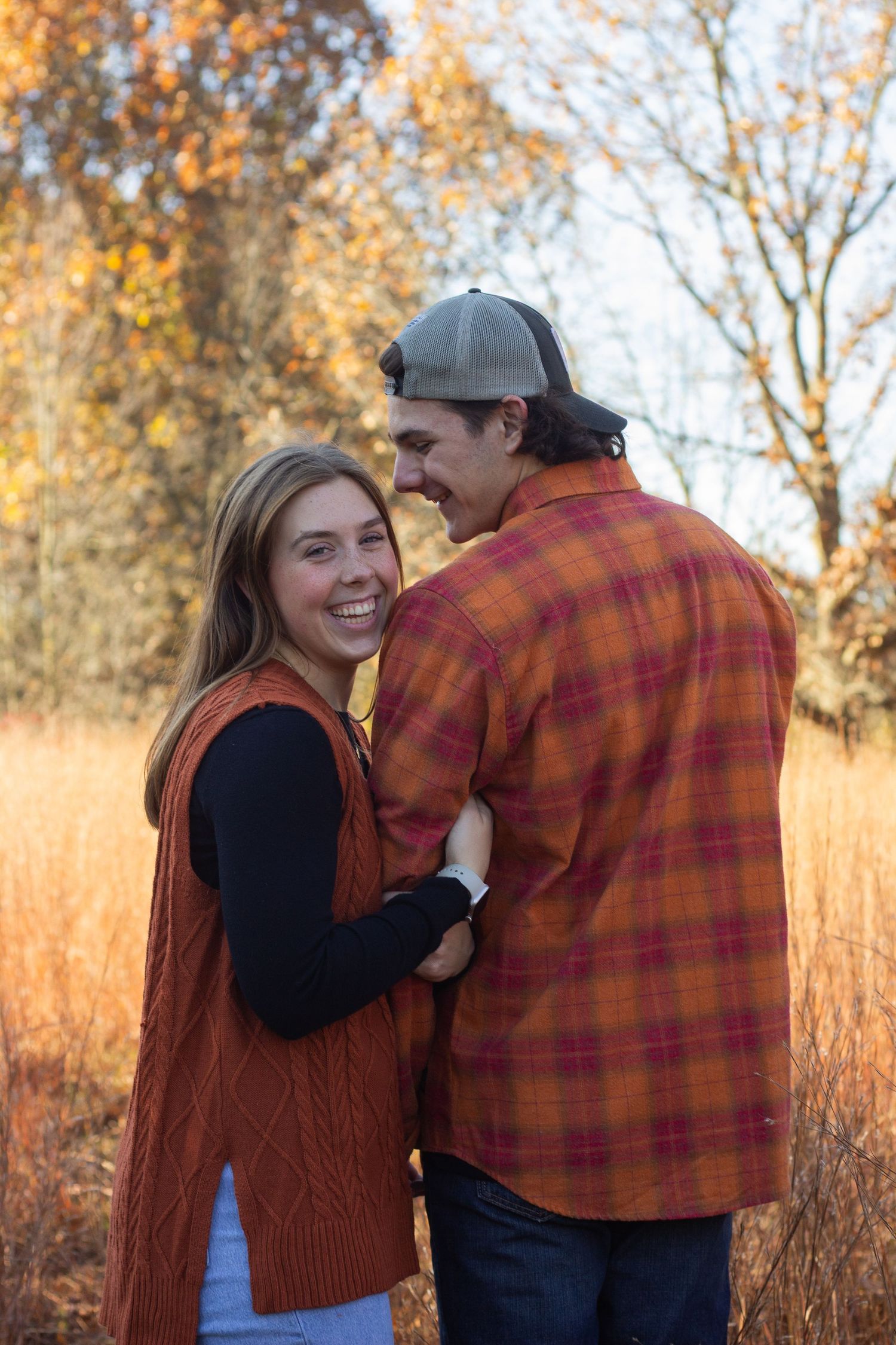 A couple shares a tender moment in a golden autumn field with tall grass and trees in warm evening light.