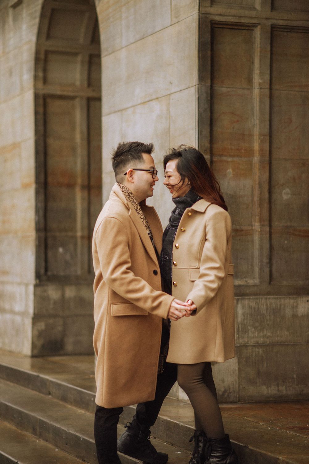 A romantic couple in matching camel coats share an intimate moment against a stone architectural backdrop.