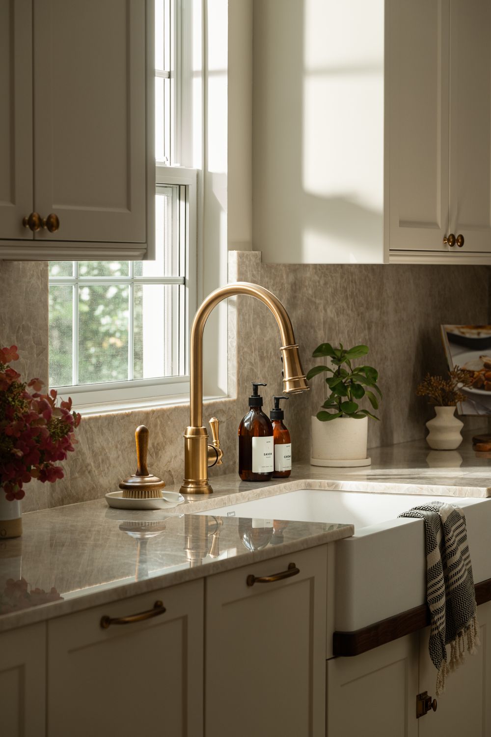 Elegant kitchen with brass faucet, marble countertops, white cabinets, farmhouse sink, and natural light from window.