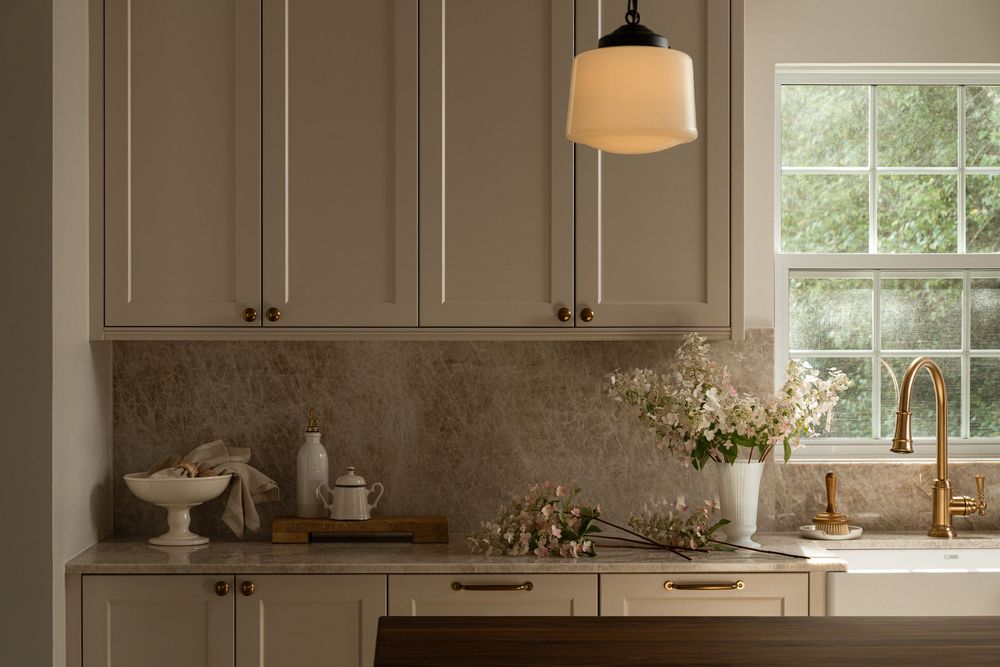 Elegant kitchen with cream cabinets, marble backsplash, brass faucet, pendant light, and fresh flowers by a window.