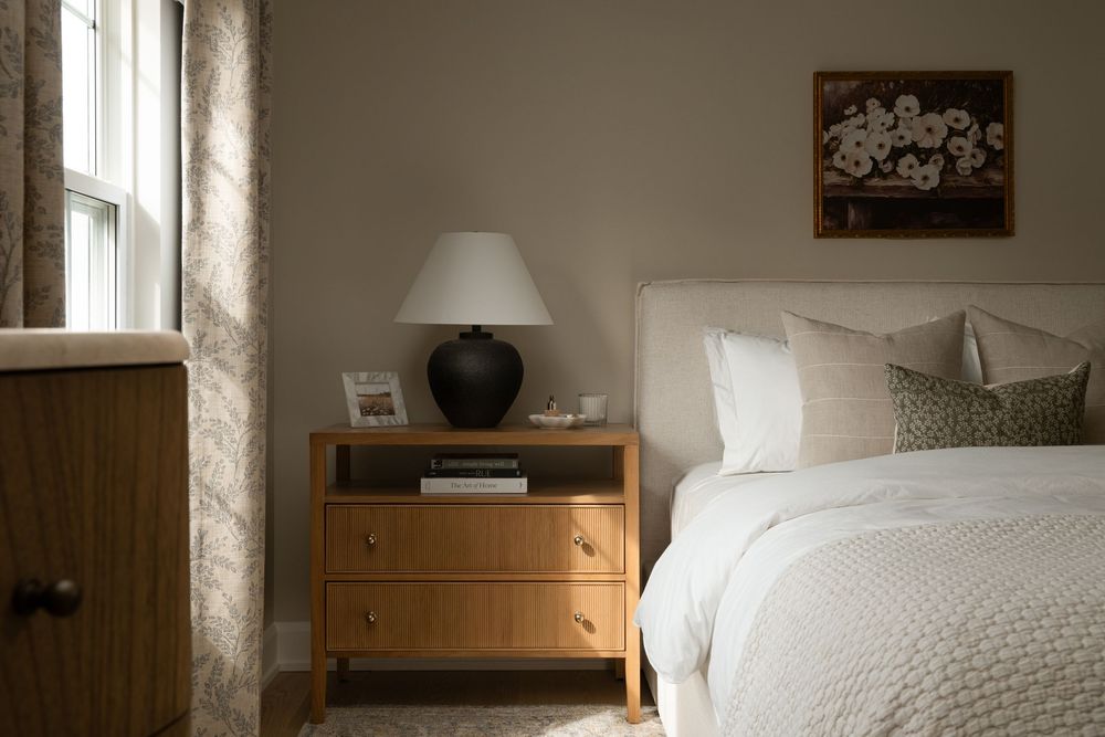 Cozy bedroom with wooden nightstand, black lamp, white bedding, floral artwork, and neutral walls near a window.