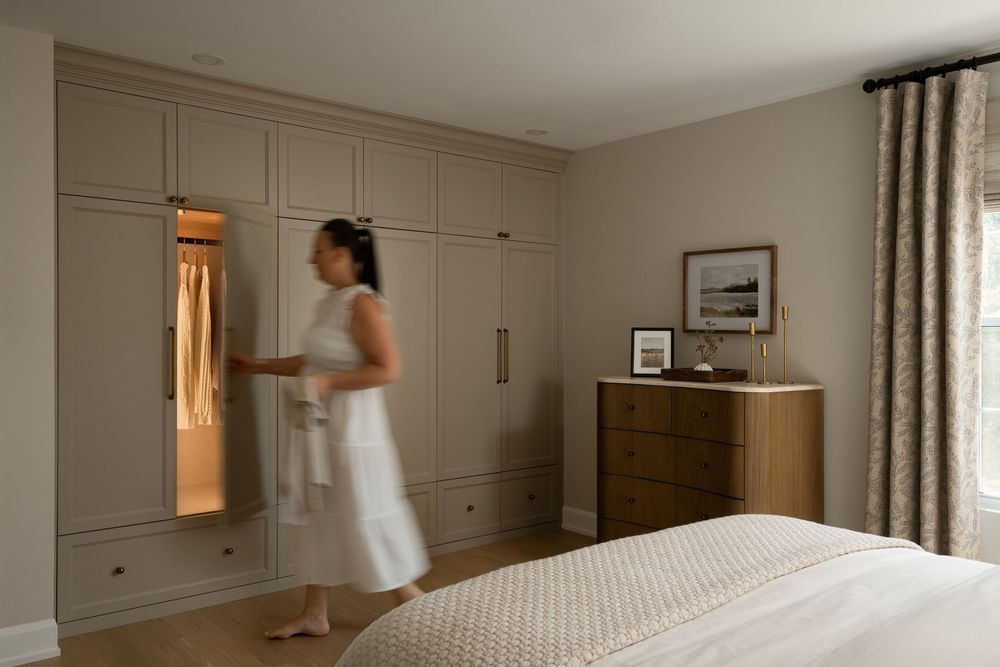 Woman in white dress opening built-in wardrobe with interior lighting in a neutral-toned bedroom.