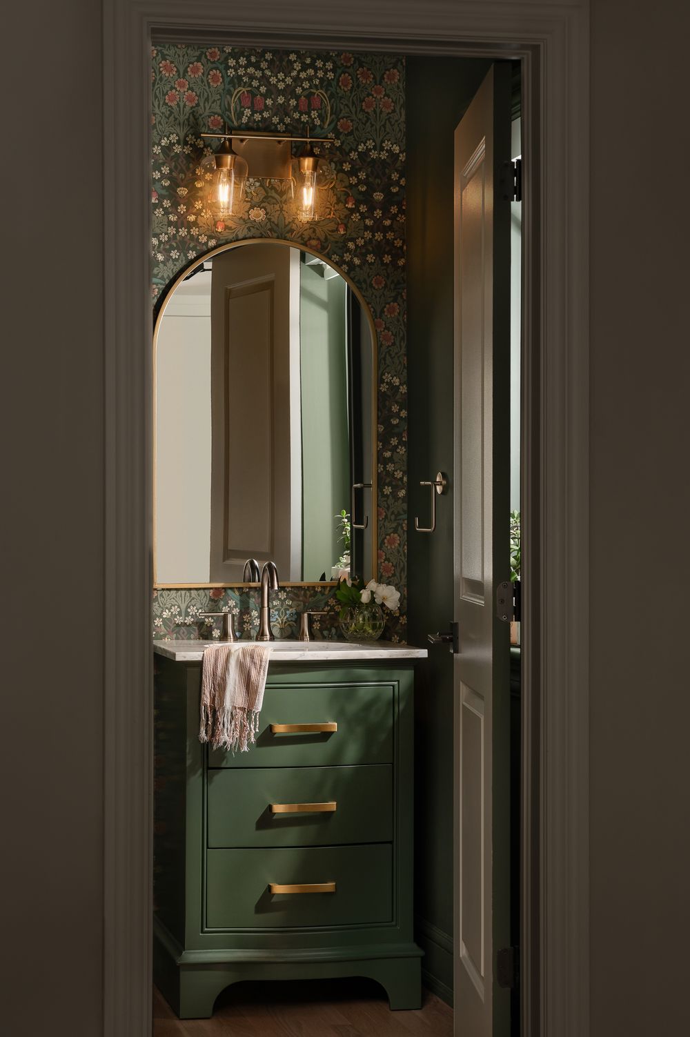 A modern bathroom vanity in dark green with brass hardware and floral wallpaper features pendant lighting and a mirror.
