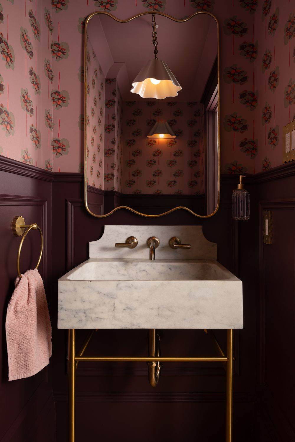 Elegant bathroom vanity with marble sink, brass fixtures, vintage pendant light, and pink floral wallpaper above dark wood paneling.