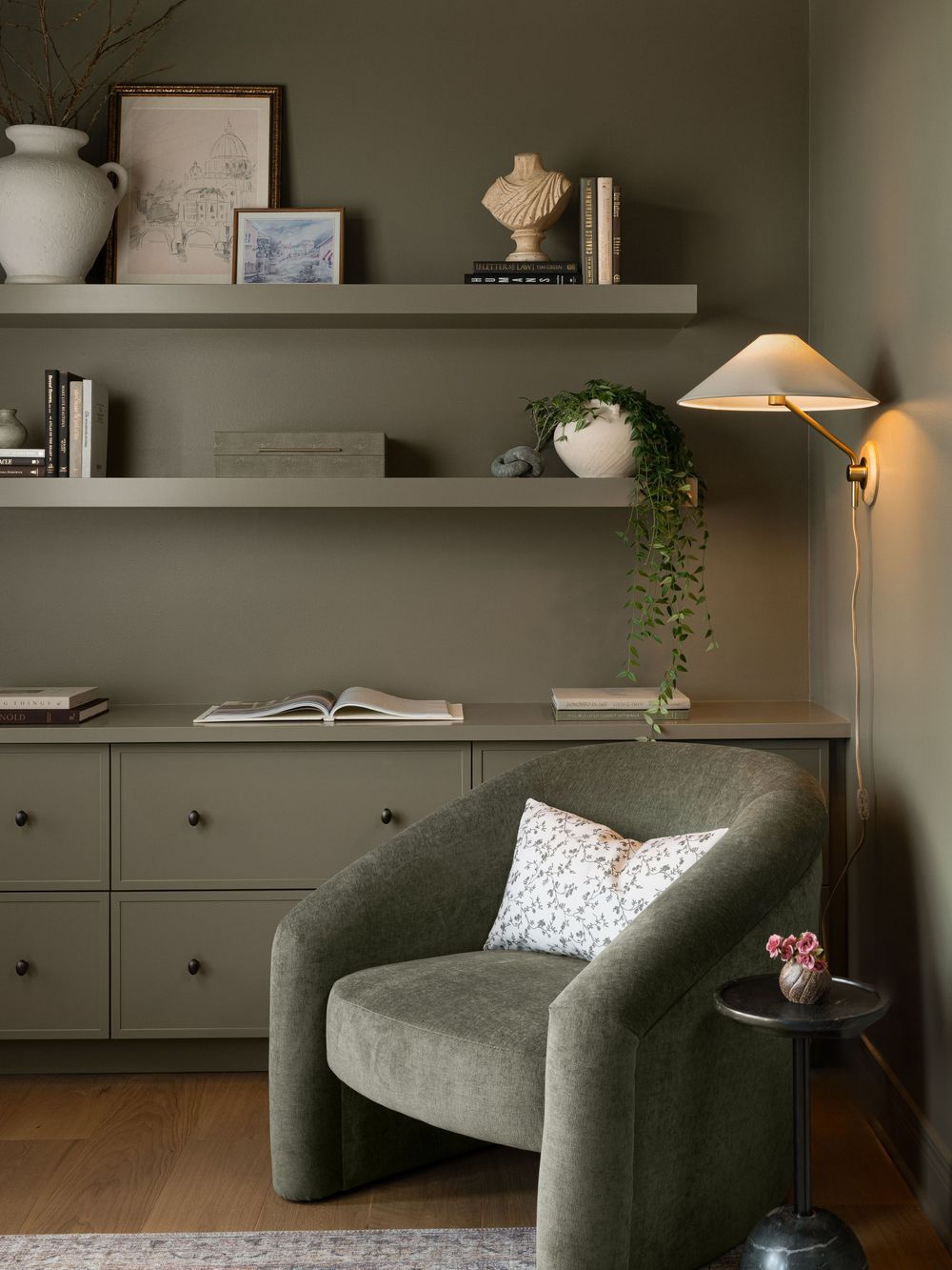 Cozy reading nook with olive green armchair, matching built-in drawers, floating shelves with decor, and a brass wall lamp.