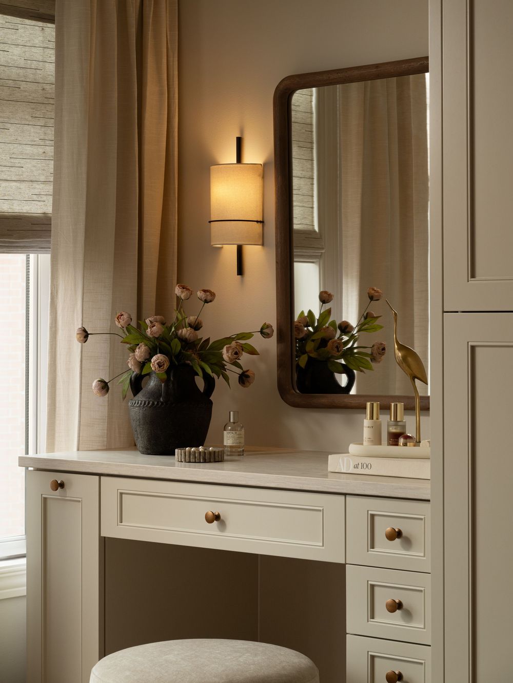 Elegant vanity desk with cream cabinetry, arched mirror, wall sconce, and floral arrangement in a serene neutral bedroom.