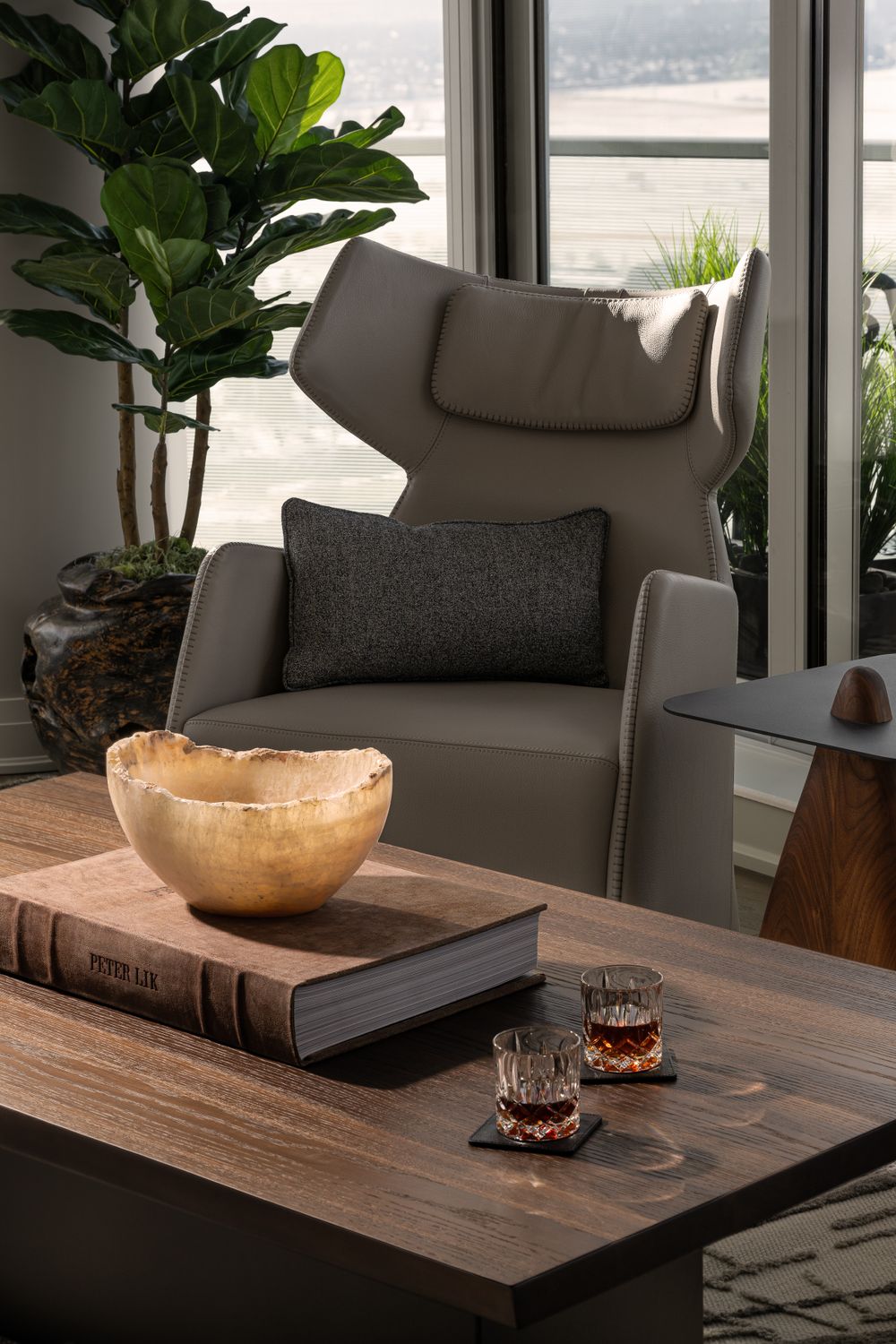 A cozy modern living room corner with gray armchair, fiddle leaf fig plant, and wooden coffee table displaying a golden bowl.