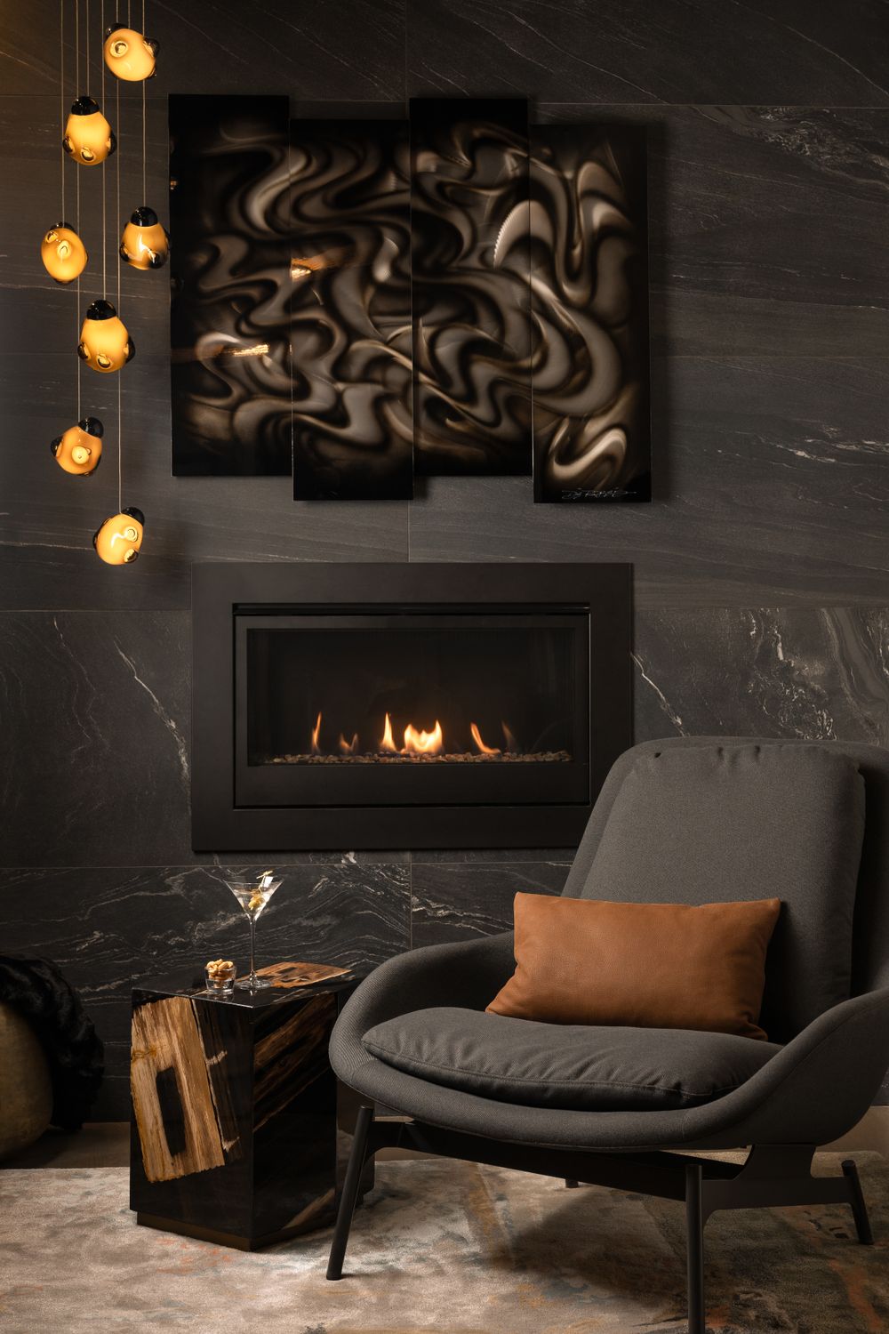 Modern living room with fireplace, gray lounge chair, brown pillow, pendant lights and abstract wall art on dark marble wall.