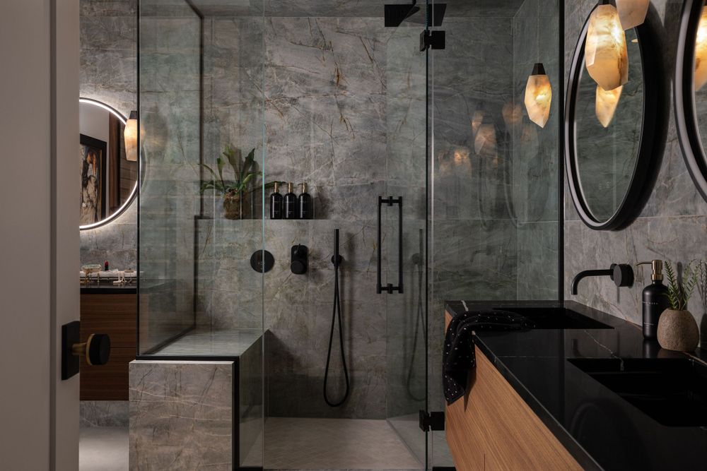 Modern luxury bathroom with marble walls, glass shower enclosure, black fixtures, and pendant lighting.