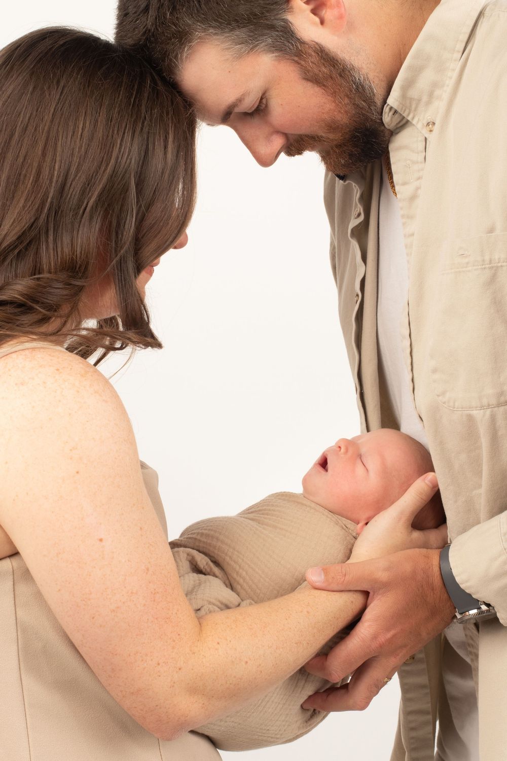 Newborn session in the studio. Mom and dad are leaning over baby with a clean white background.