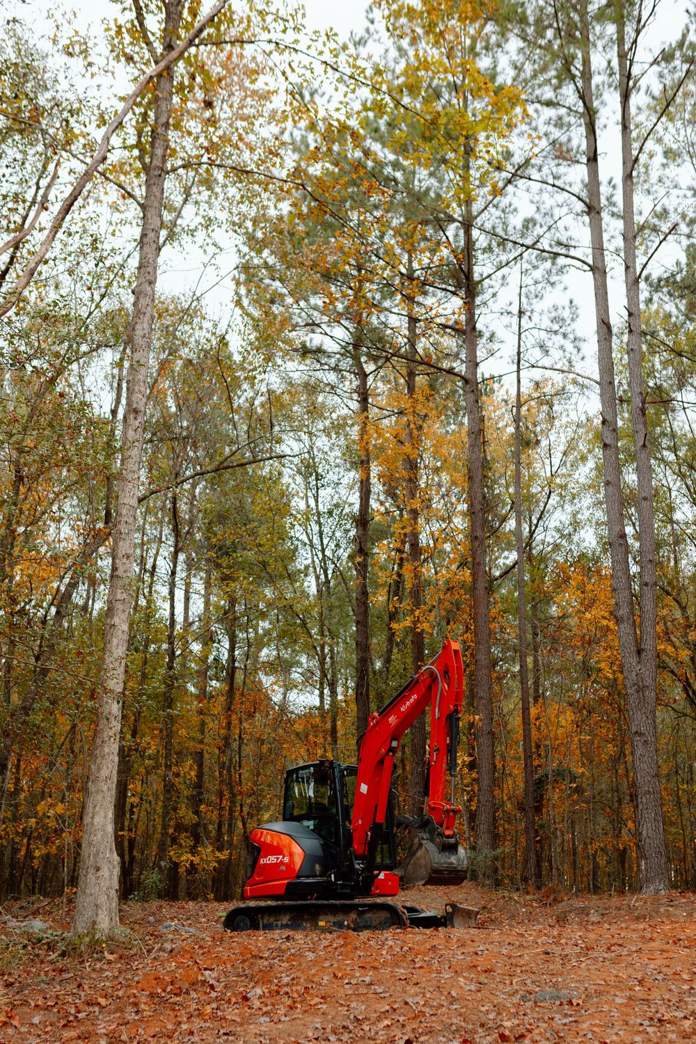 Red Kubota mini excavator parked in an autumn forest with colorful foliage.