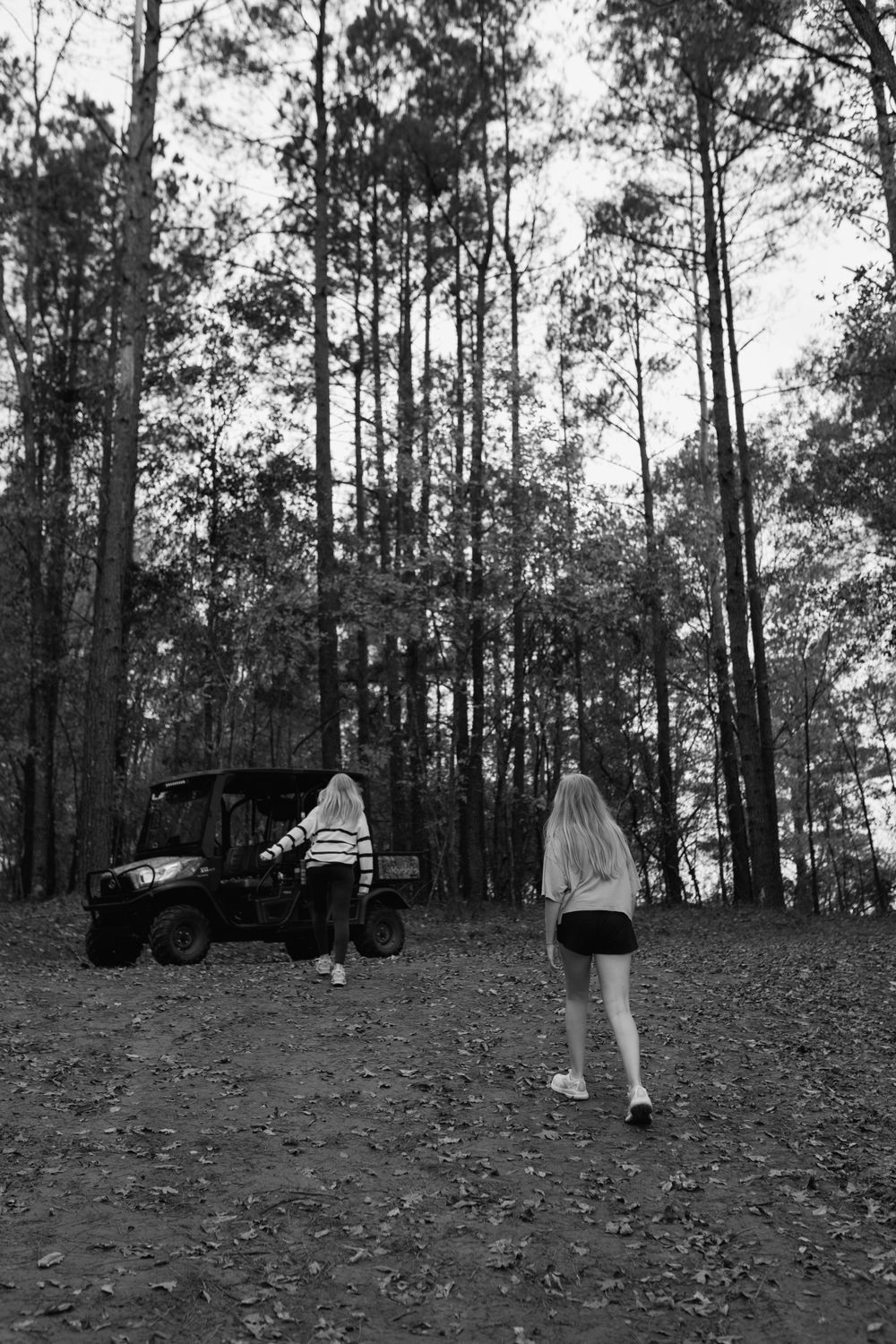 Two young women walk toward an ATV parked in a tall pine forest.