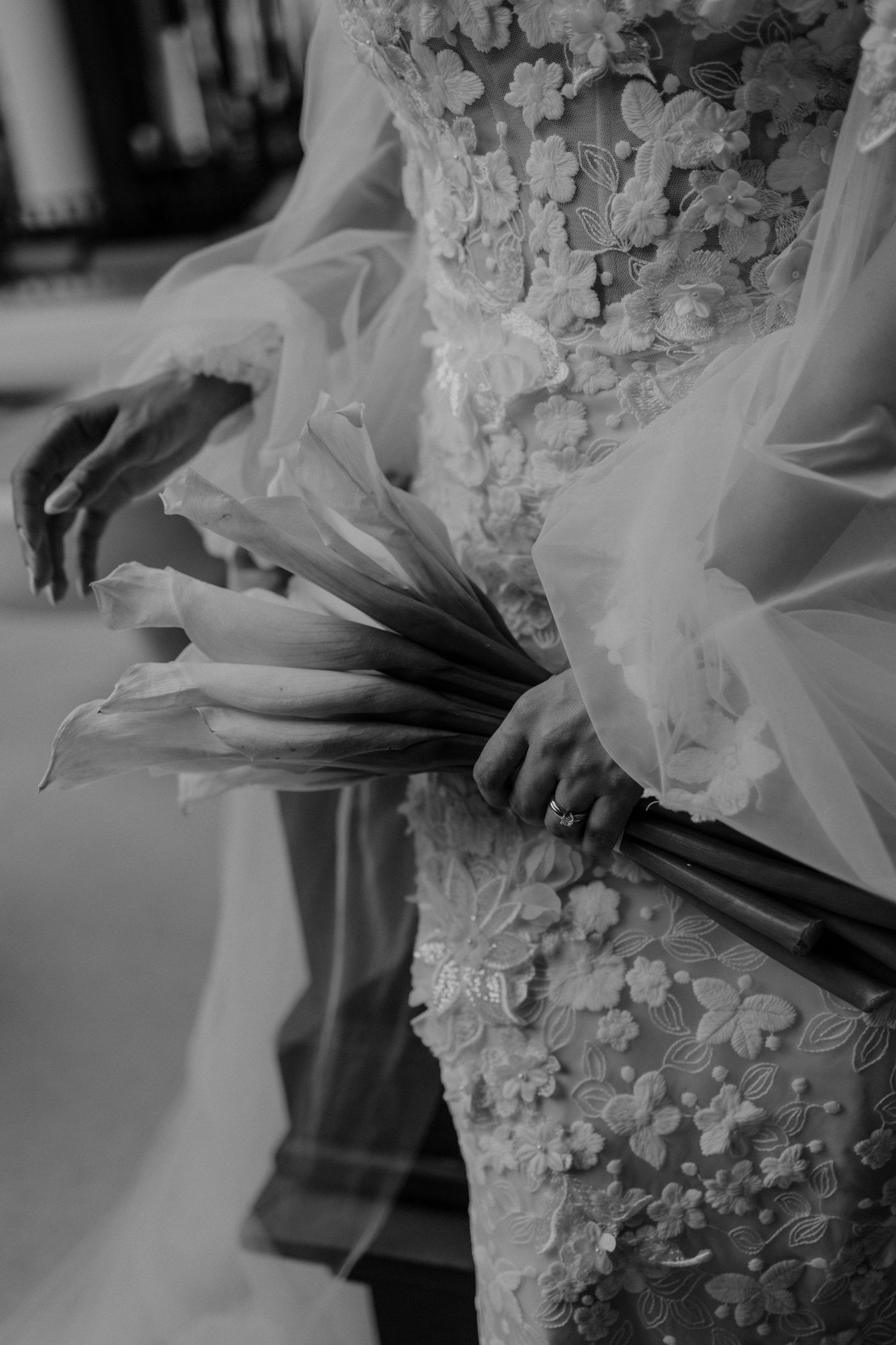 Black and white photo of a bride holding calla lily bouquet, wearing floral lace wedding gown with sheer sleeves.