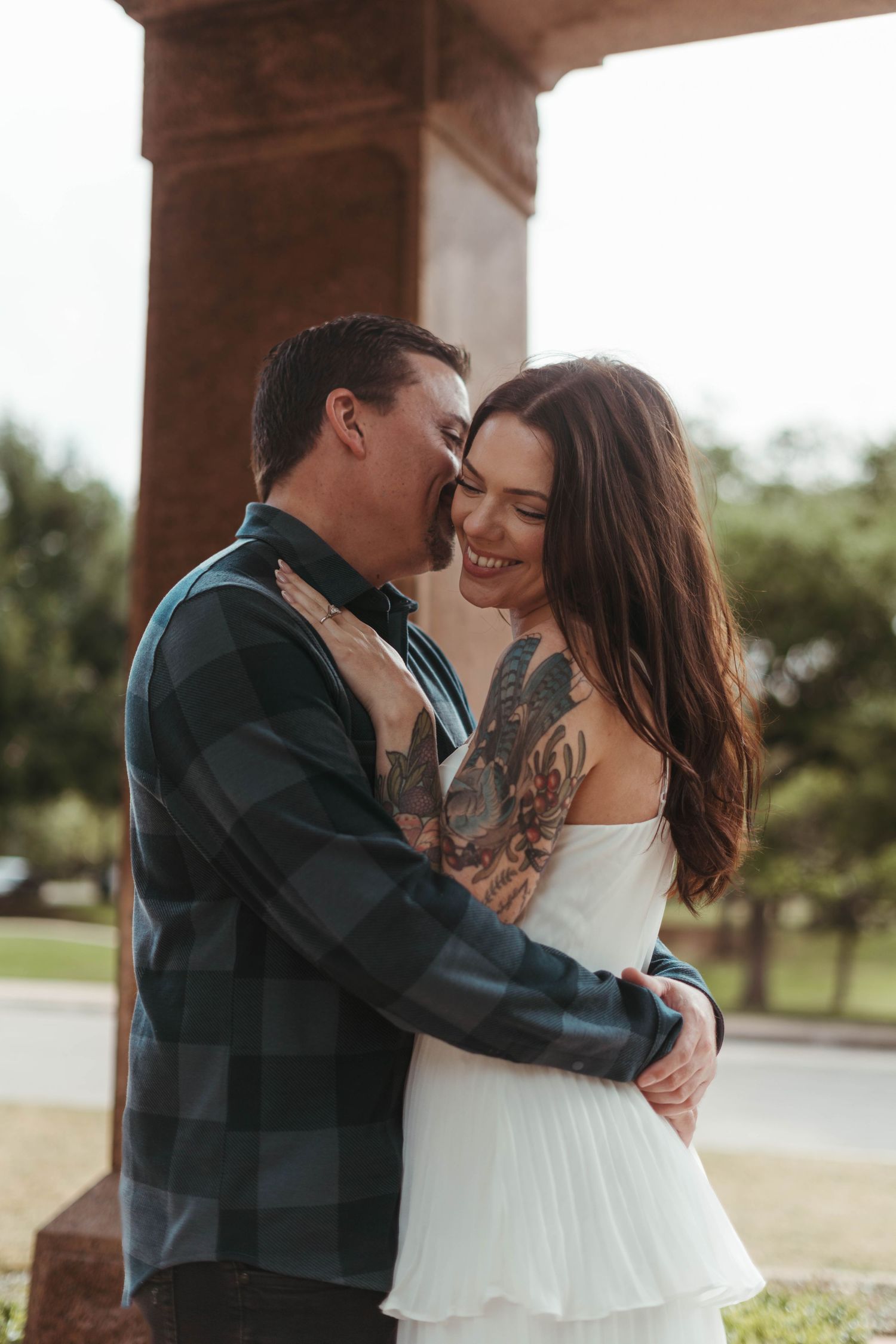 A couple embraces intimately outdoors, with one wearing a plaid shirt and the other in a flowing white dress.