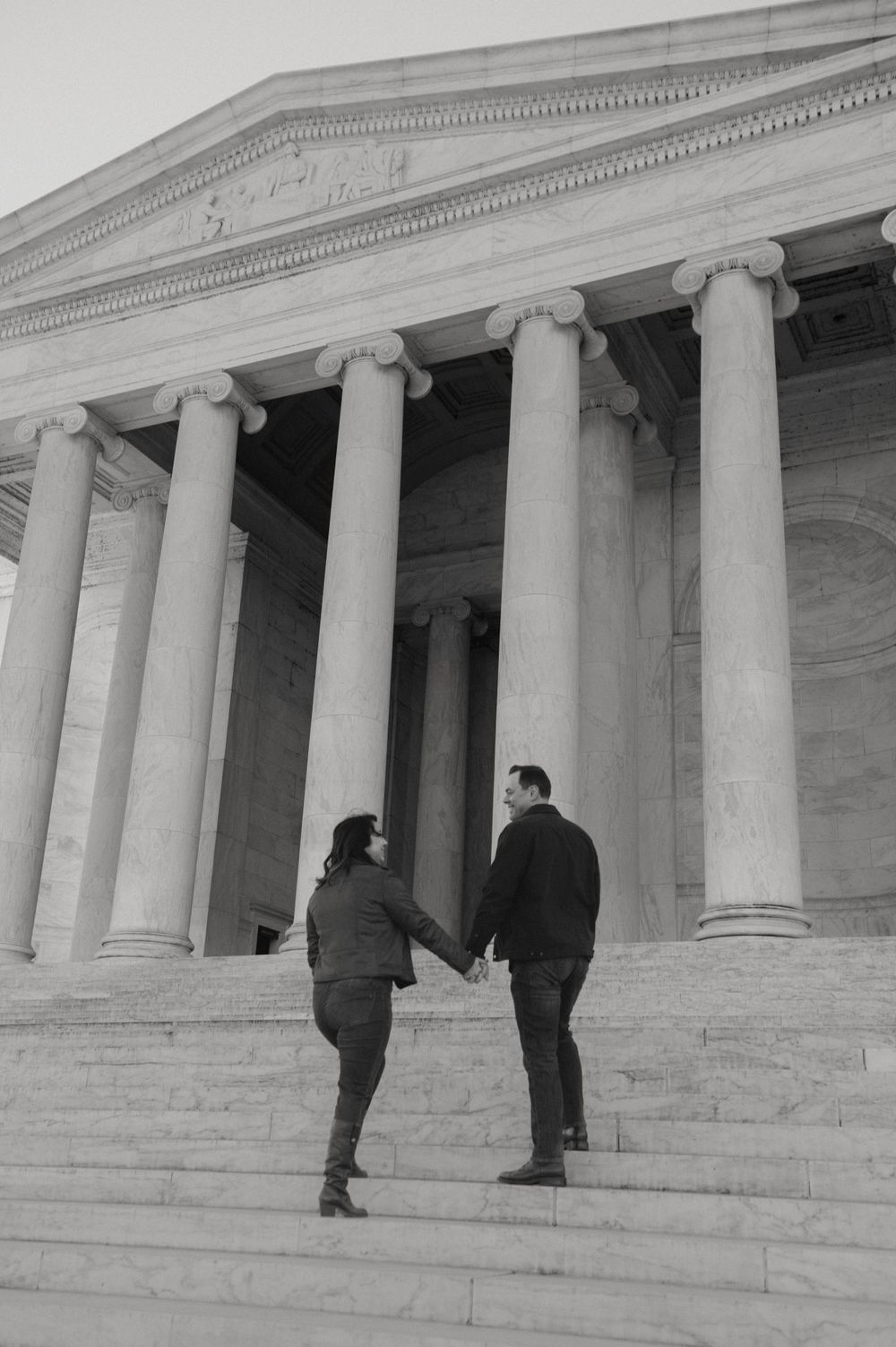 A couple holds hands while climbing the steps of a neoclassical building with white columns.