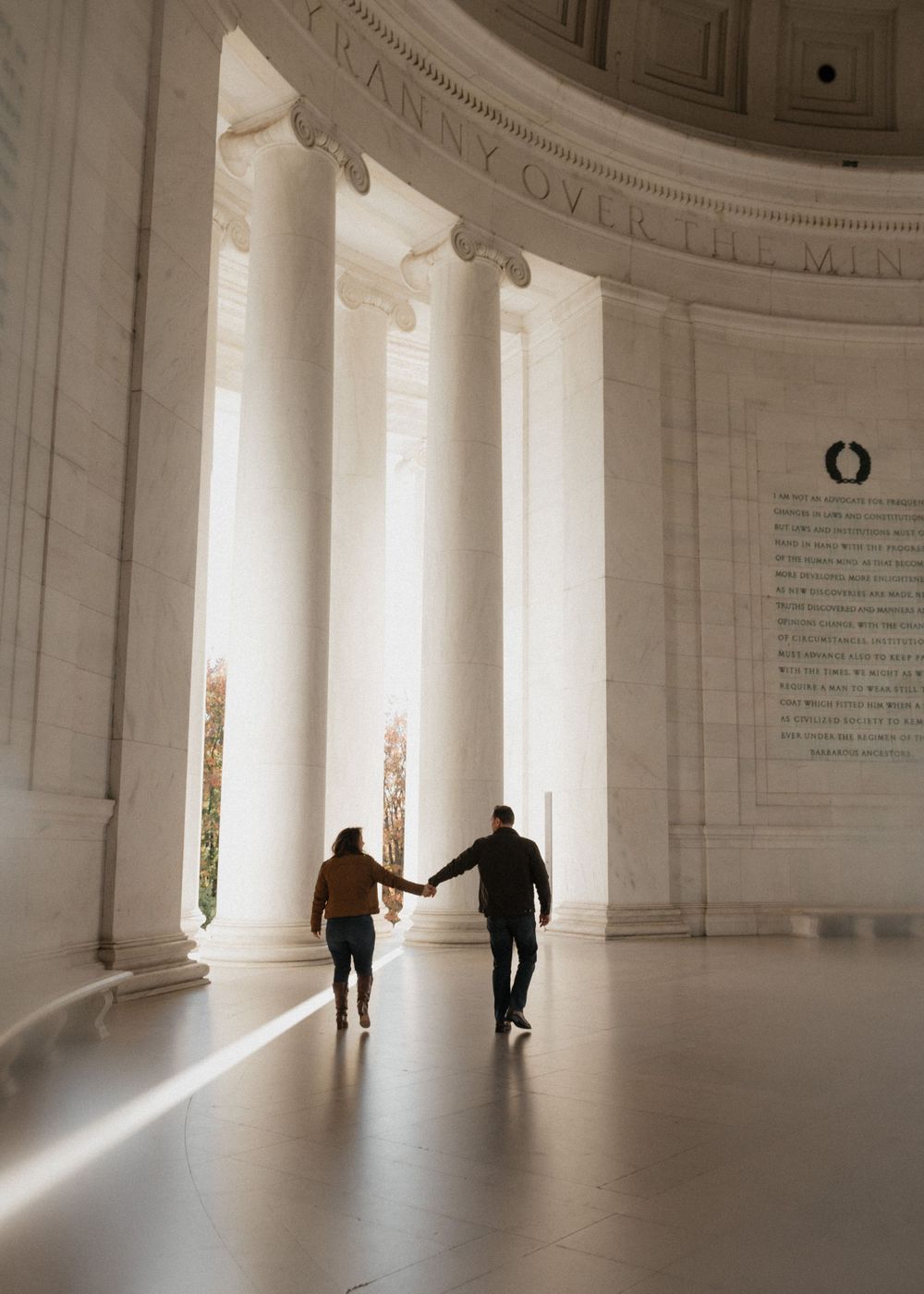 Couple holding hands while walking through white marble colonnade with sunlight streaming in.