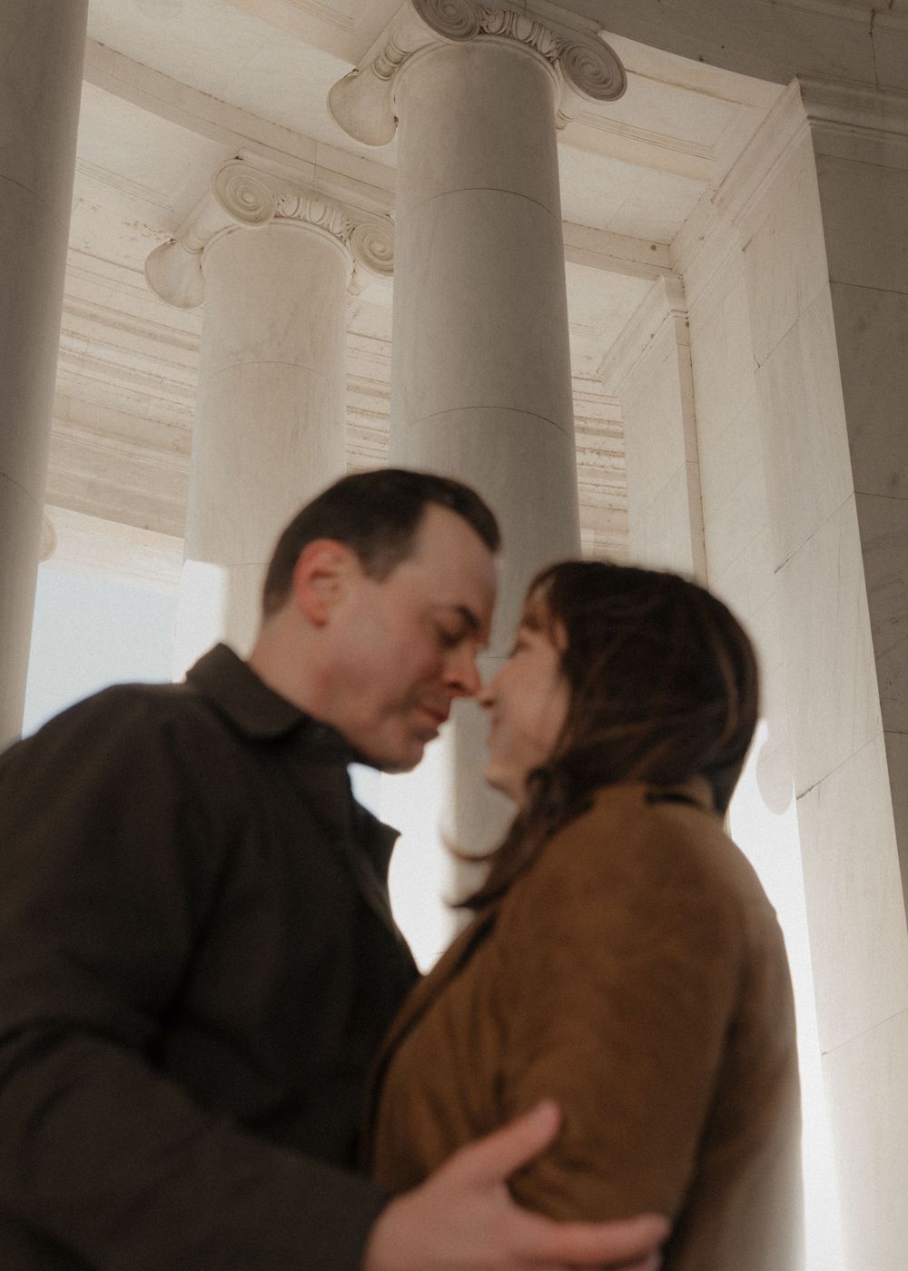 Tender moment shared between couple in brown jackets against white architectural pillars.