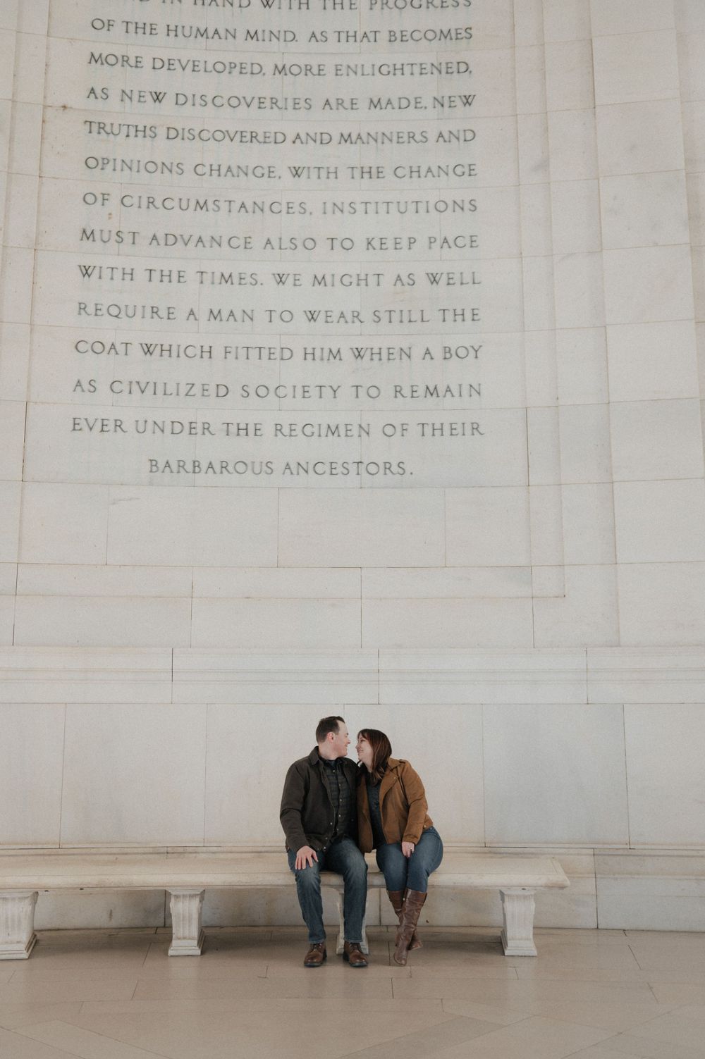 Couple sitting on bench beneath carved text on marble wall in memorial building.