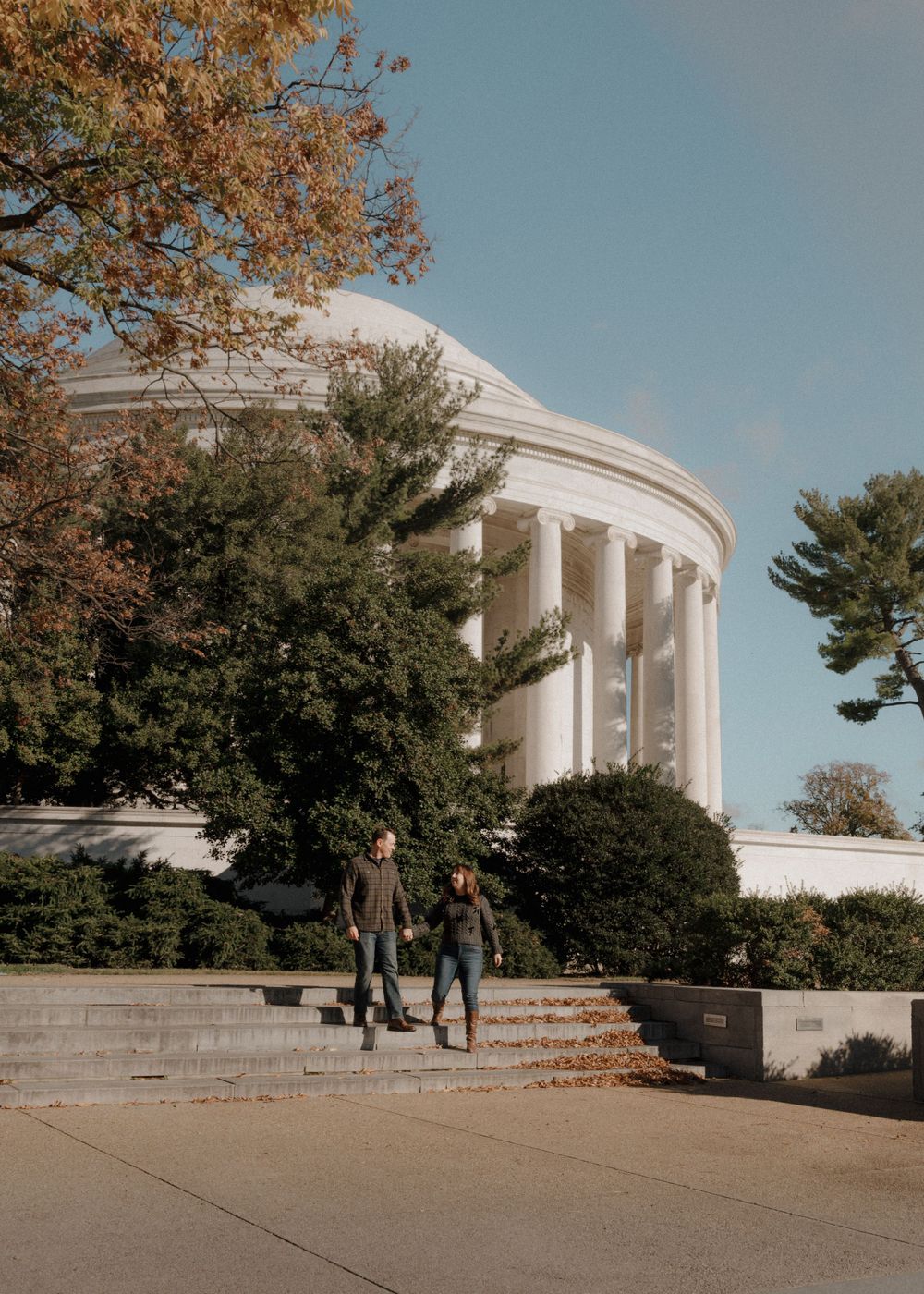 Couple walking near Jefferson Memorial with autumn trees and white columns in view.