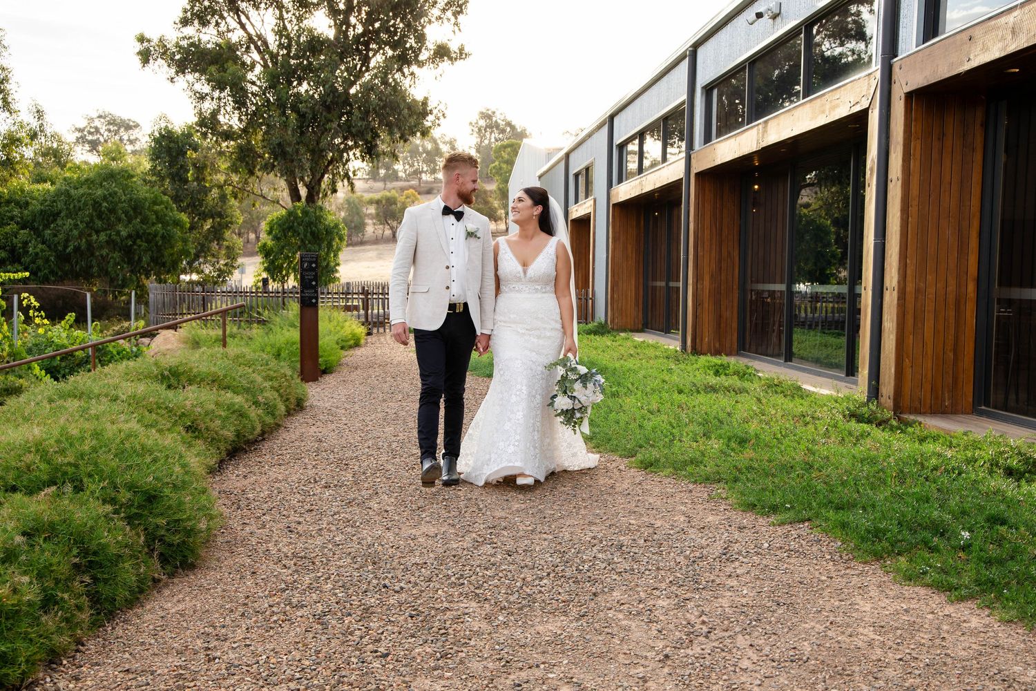 A couple walks along a gravel path beside modern architecture and landscaped gardens.