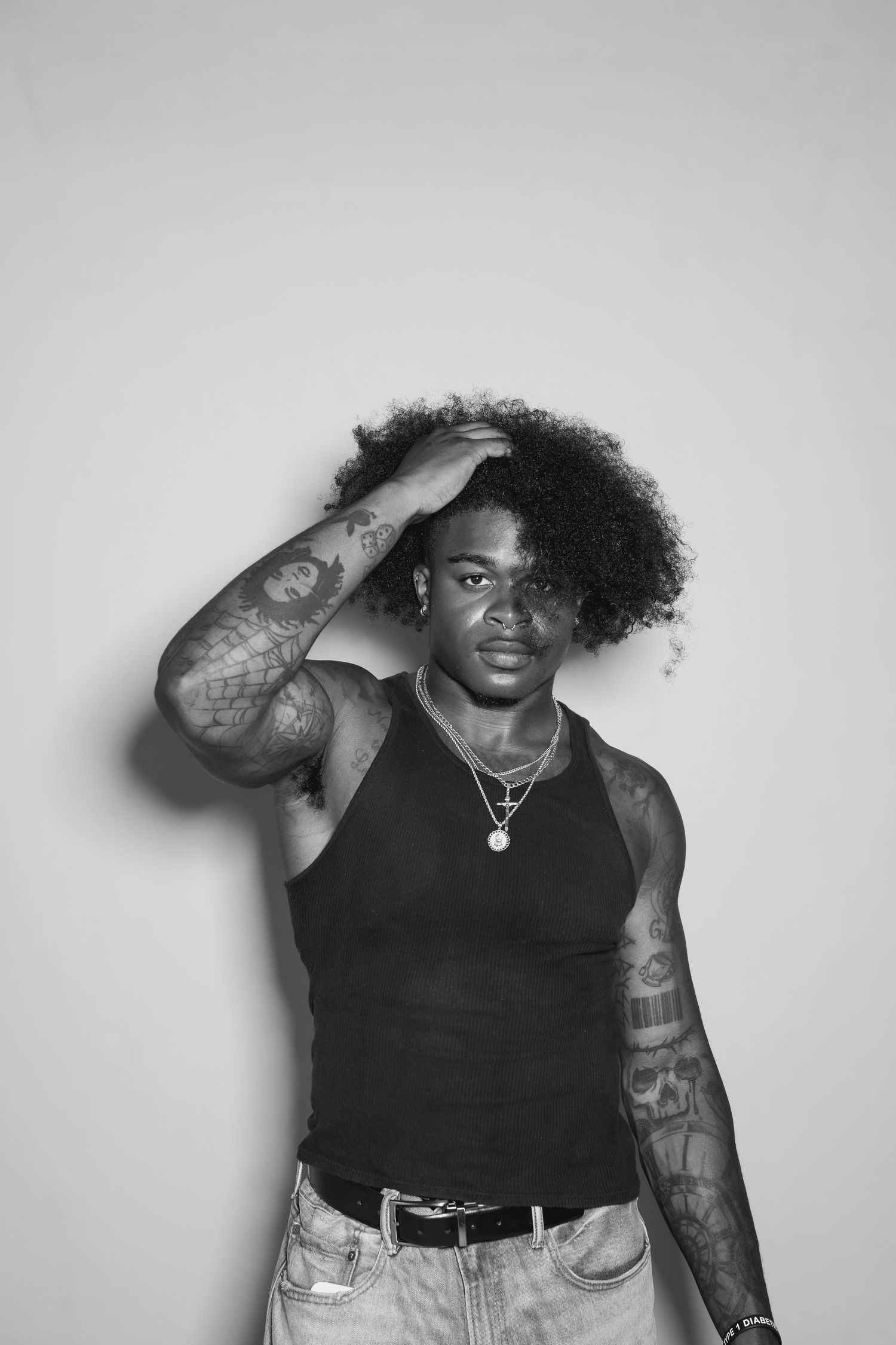 A black and white portrait of someone in a tank top showing off tattooed arms against a plain background.
