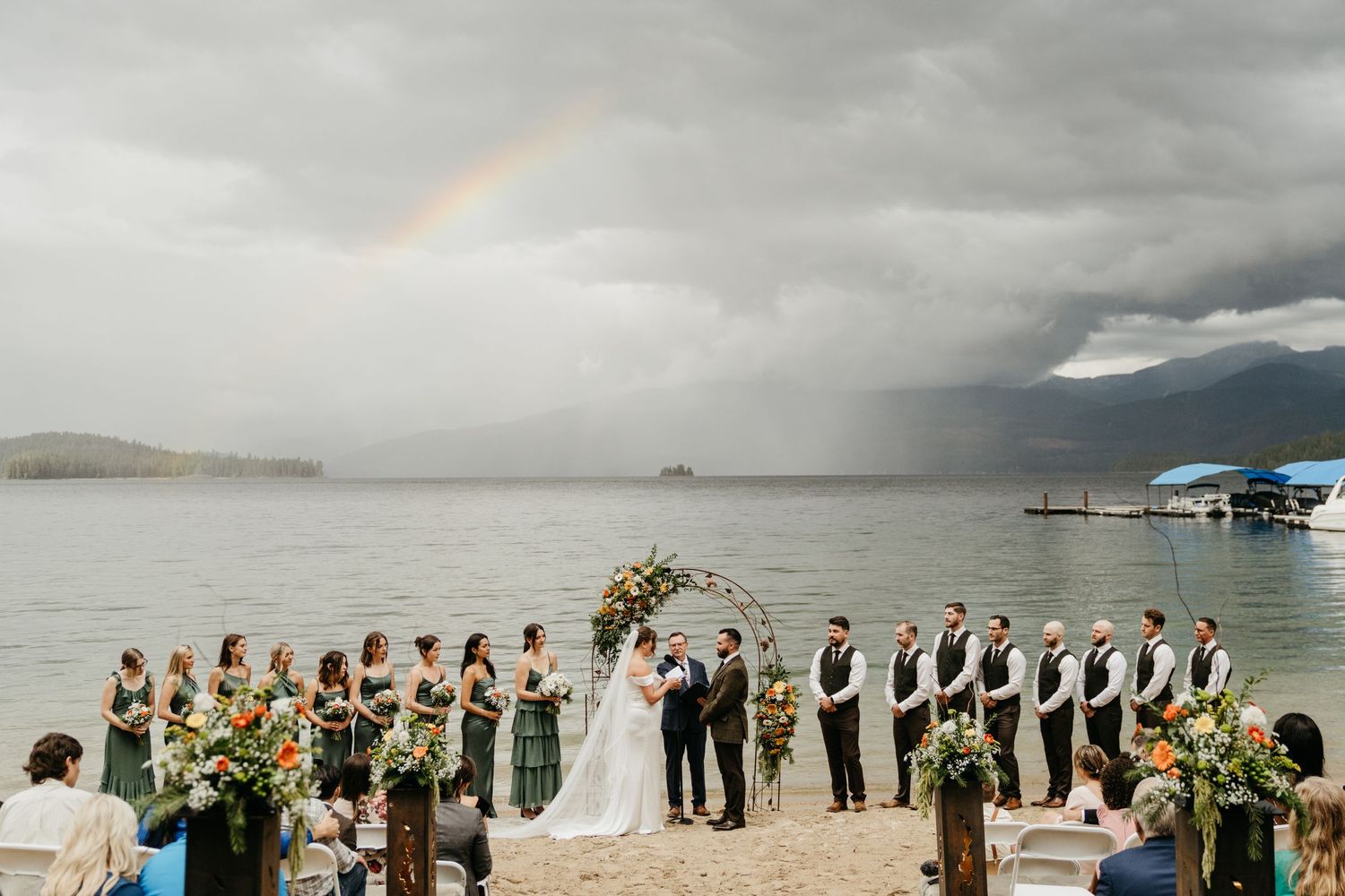 Wedding ceremony on beach with dramatic stormy sky and rainbow overhead during lakeside celebration.