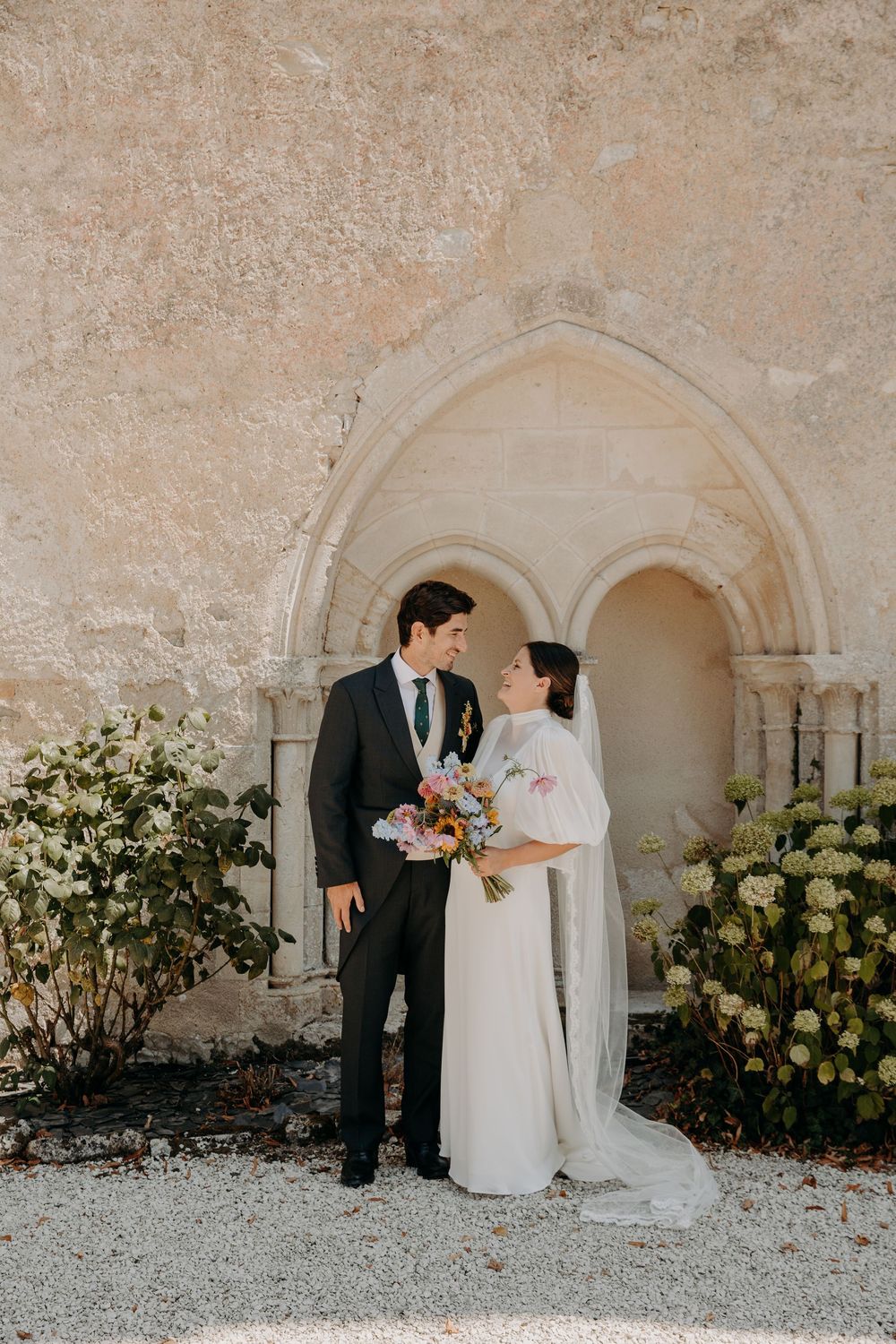 Bride and groom share a romantic moment in front of a stone Gothic arch, bride holding a colorful bouquet.