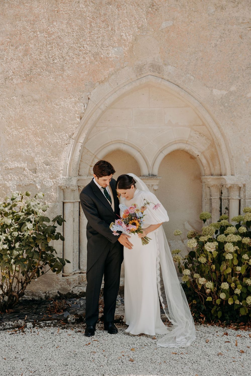 Bride and groom stand together before a stone Gothic arch, bride holding a colorful bouquet in a white gown with veil.
