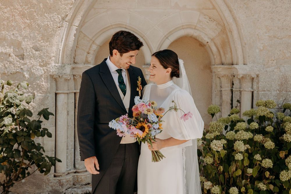 Bride and groom smile at each other outside a stone church, bride holding a colorful wildflower bouquet in a white dress.