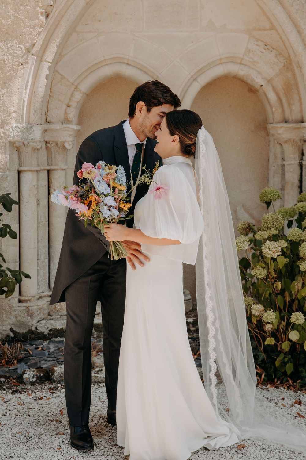 A bride and groom share a romantic moment, holding a colorful bouquet, in front of a stone archway outdoors.