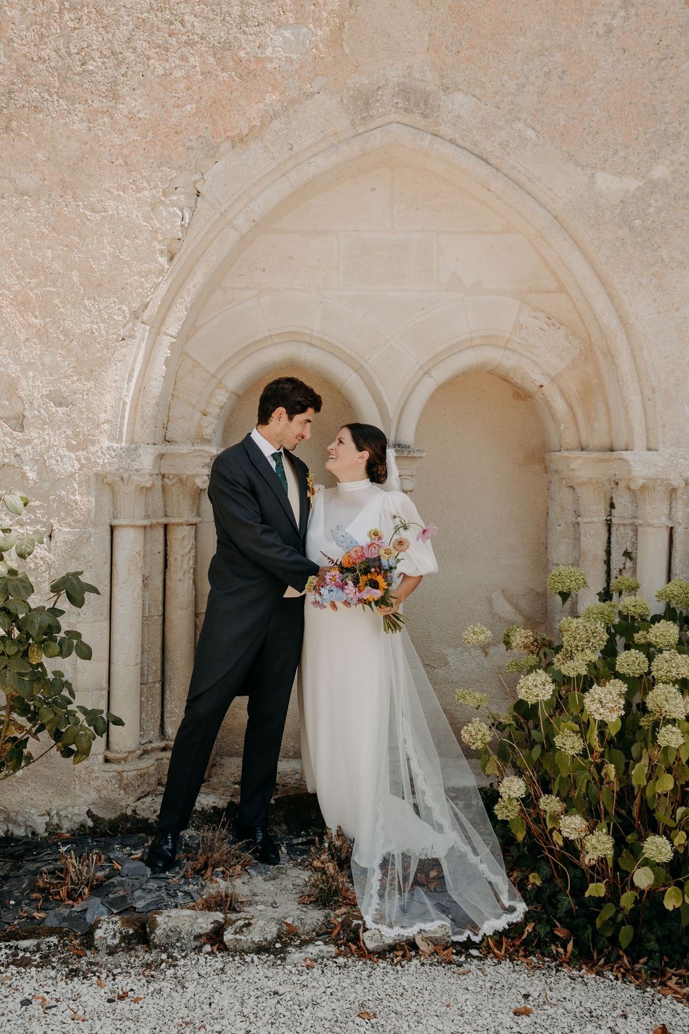 Bride and groom stand together before a stone Gothic arch, she holds a colorful bouquet, both in elegant wedding attire.