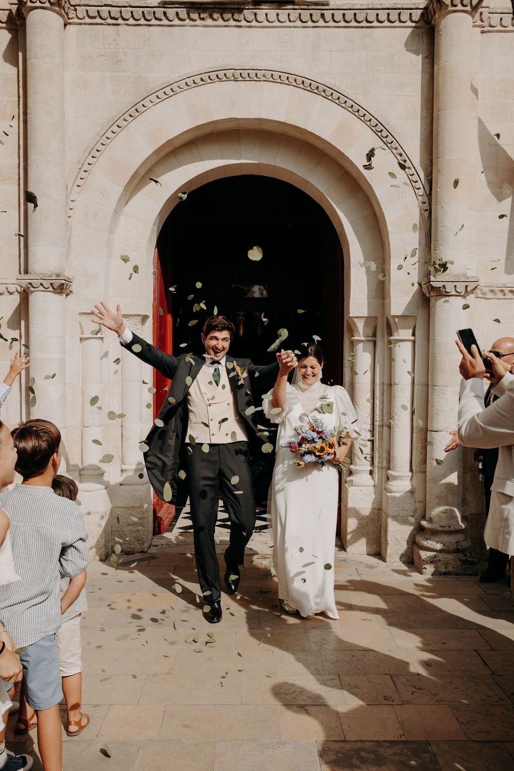 Joyful newlyweds exit church amid confetti shower, bride in white gown with bouquet, groom in black suit celebrating.