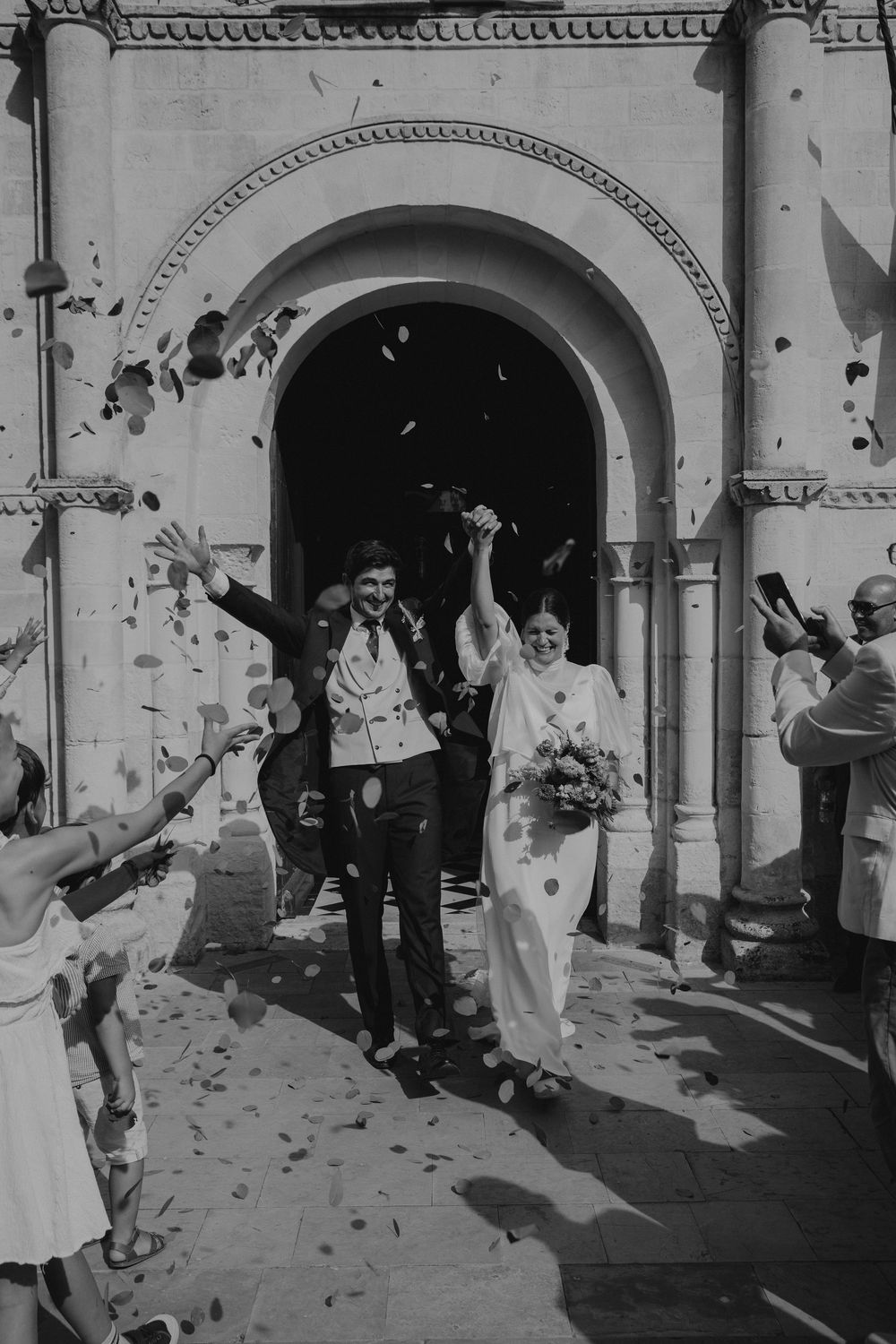 Black and white photo of a joyful couple exiting a church as wedding guests throw confetti petals.