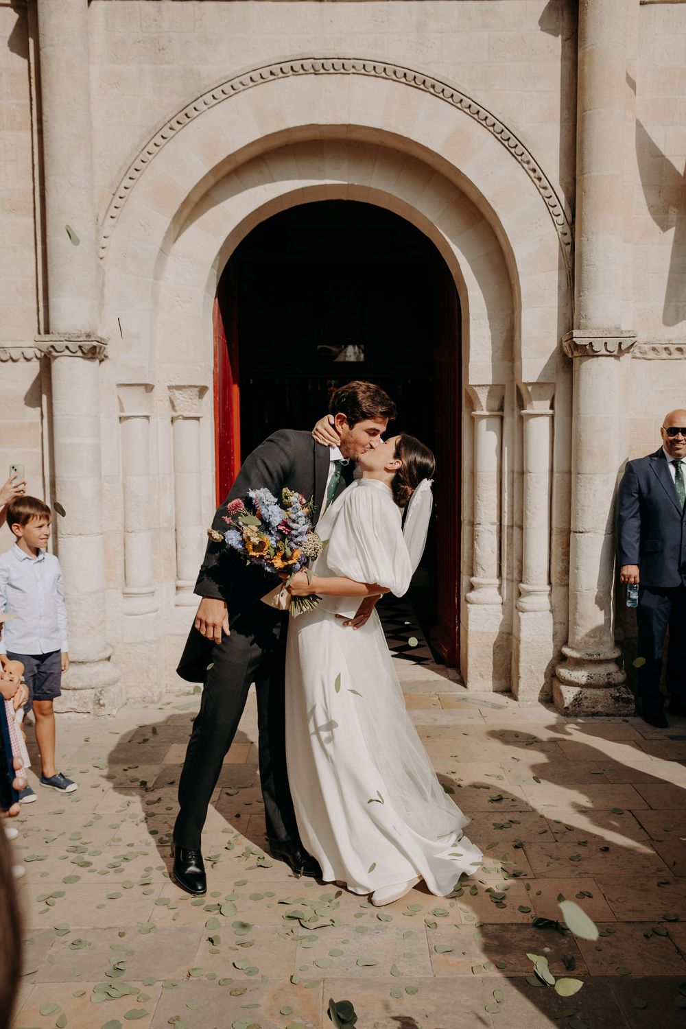 Newlywed couple sharing a first kiss outside a stone church doorway, bride in white gown holding bouquet.