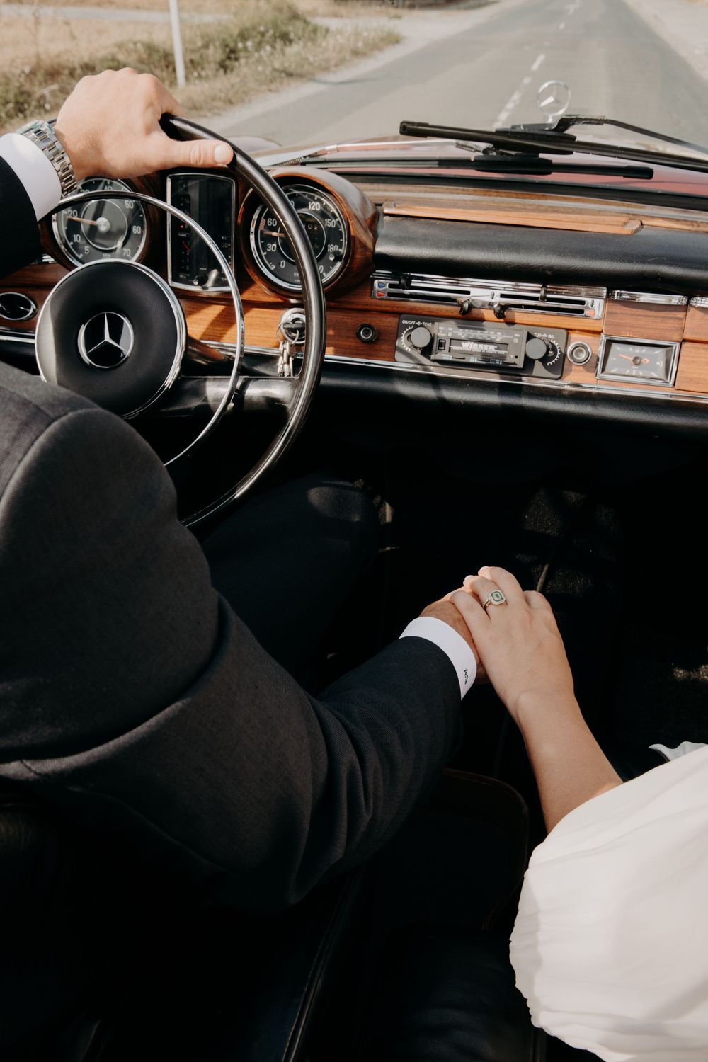 Newlyweds holding hands in a vintage Mercedes-Benz convertible, bride in white dress, groom in suit driving on scenic road.