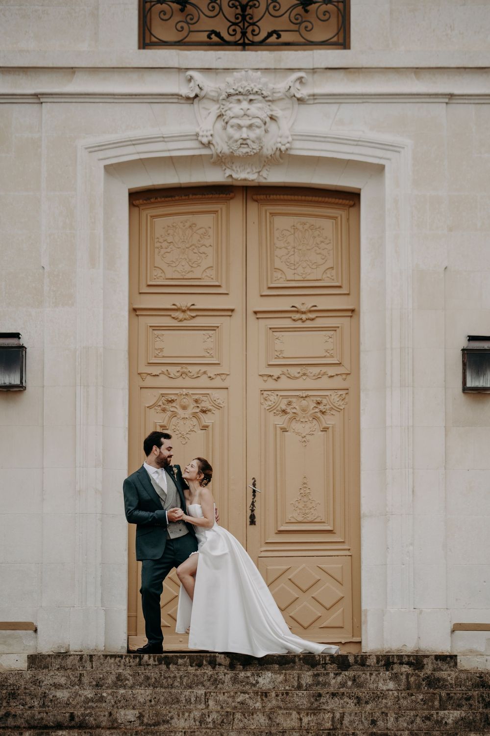 Wedding couple poses romantically against ornate cream-colored doors and stone wall.