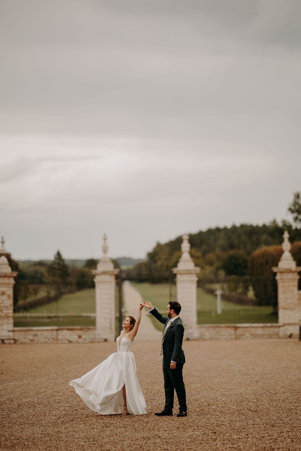 Wedding couple poses in an elegant outdoor venue with ornate stone pillars and expansive grounds.