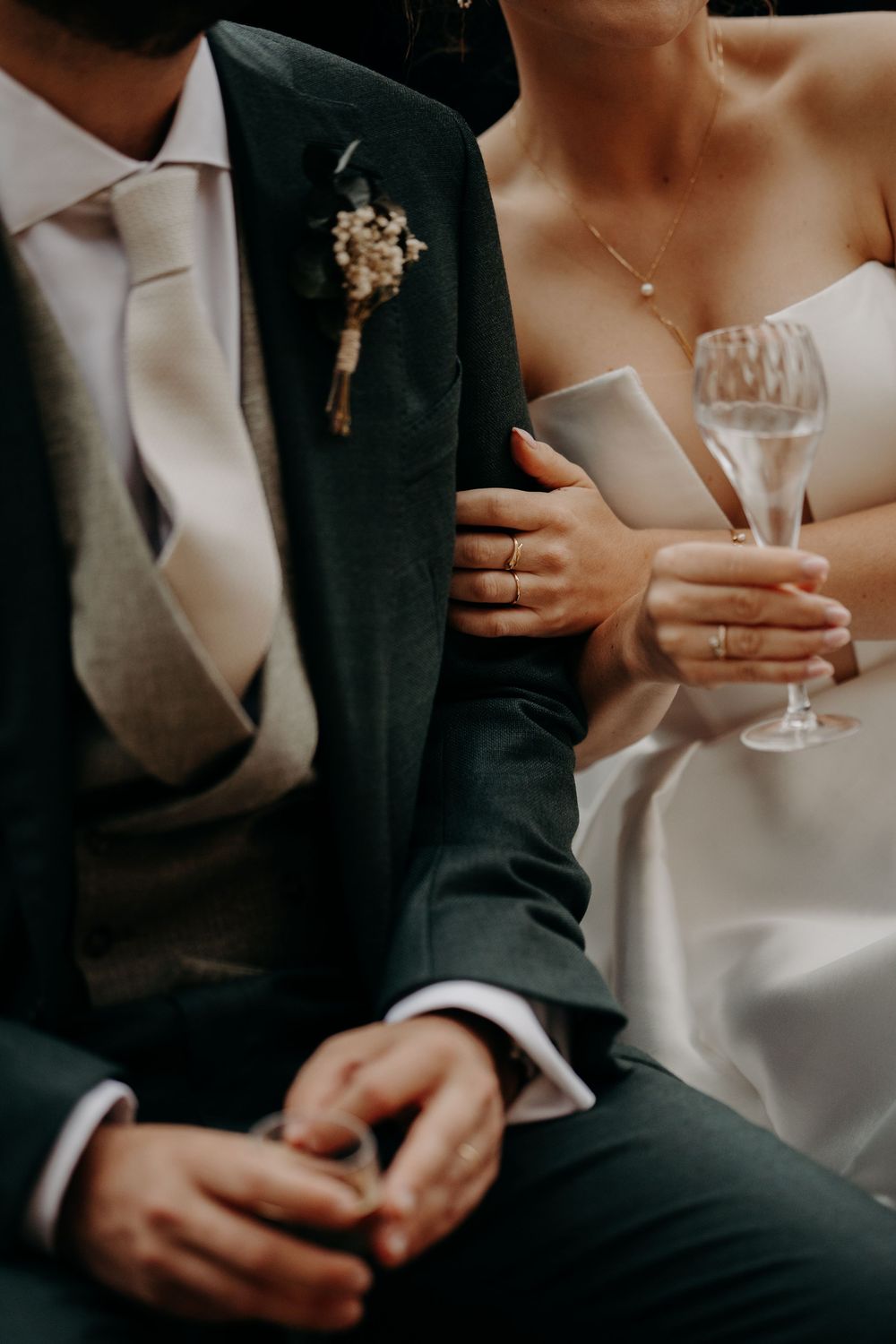 Gros plan d'une tenue de mariage formelle montrant un costume sombre avec boutonnière et une robe blanche avec un verre à champagne.