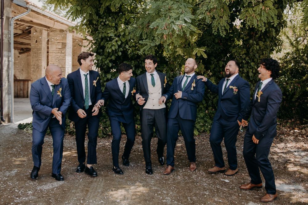 Seven groomsmen in navy suits laughing and celebrating outdoors at a wedding venue surrounded by lush greenery.