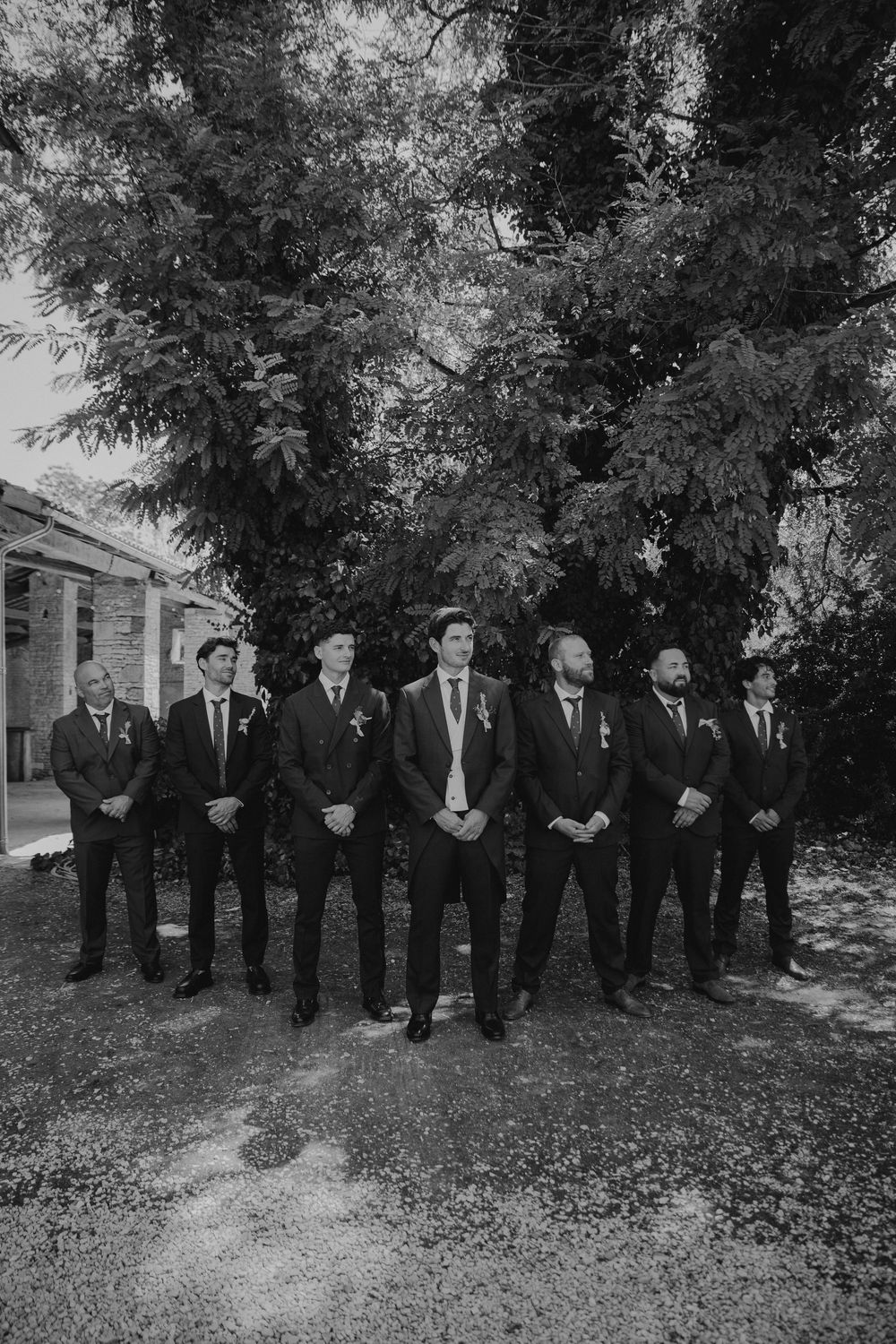 Black and white photo of a groom and groomsmen in suits standing outdoors under large trees at a wedding.
