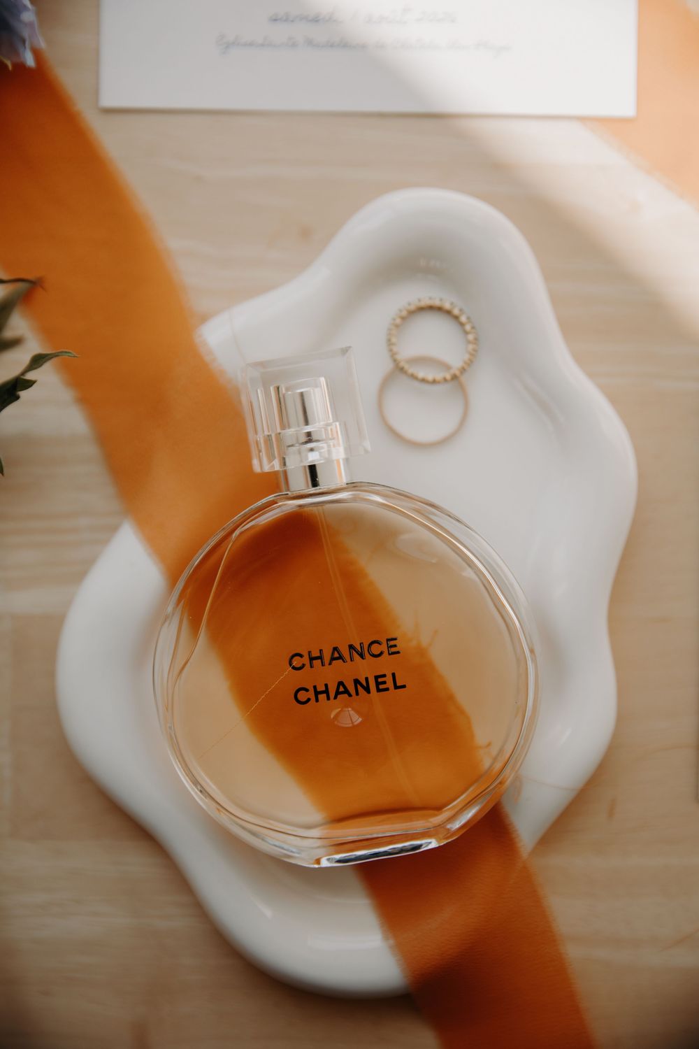 Chanel Chance perfume bottle on white ceramic tray with gold rings and orange ribbon, elegant wedding flat lay.