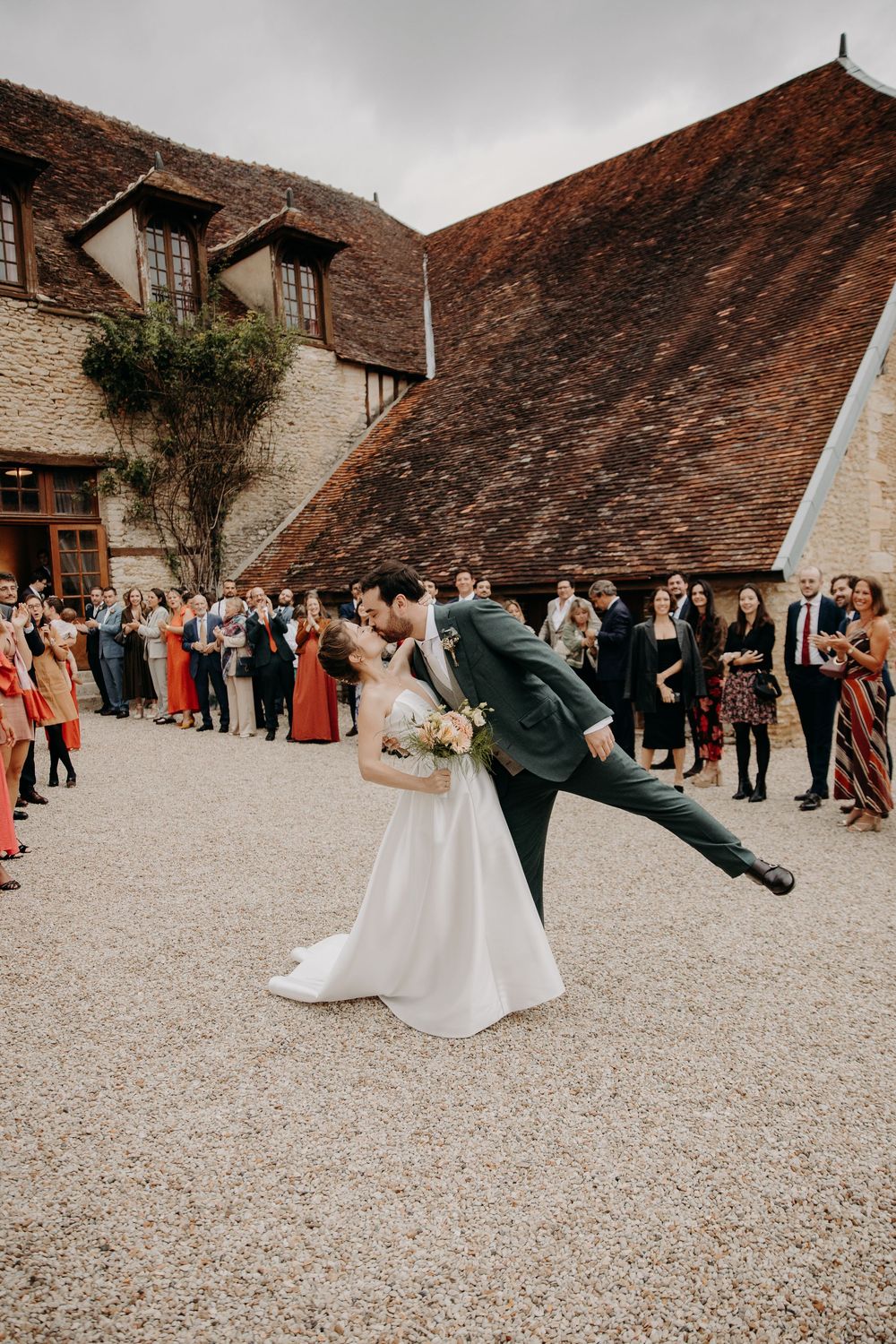 Wedding couple performs dramatic dip poses in front of historic stone building with guests watching.