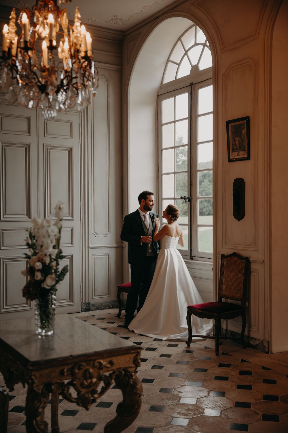 Elegant wedding couple poses in grand room with chandelier and arched windows.