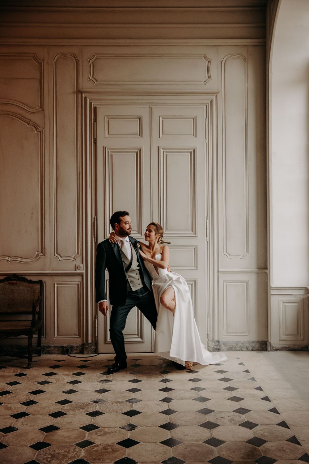Stylish wedding couple poses playfully against ornate wall panels and checkered floor.