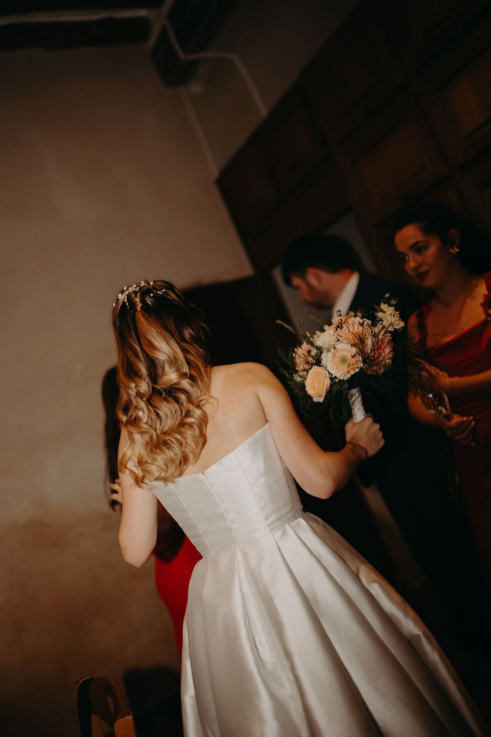 Une mariée en robe de mariée blanche danse lors d'une réception faiblement éclairée avec des invités visibles en arrière-plan.