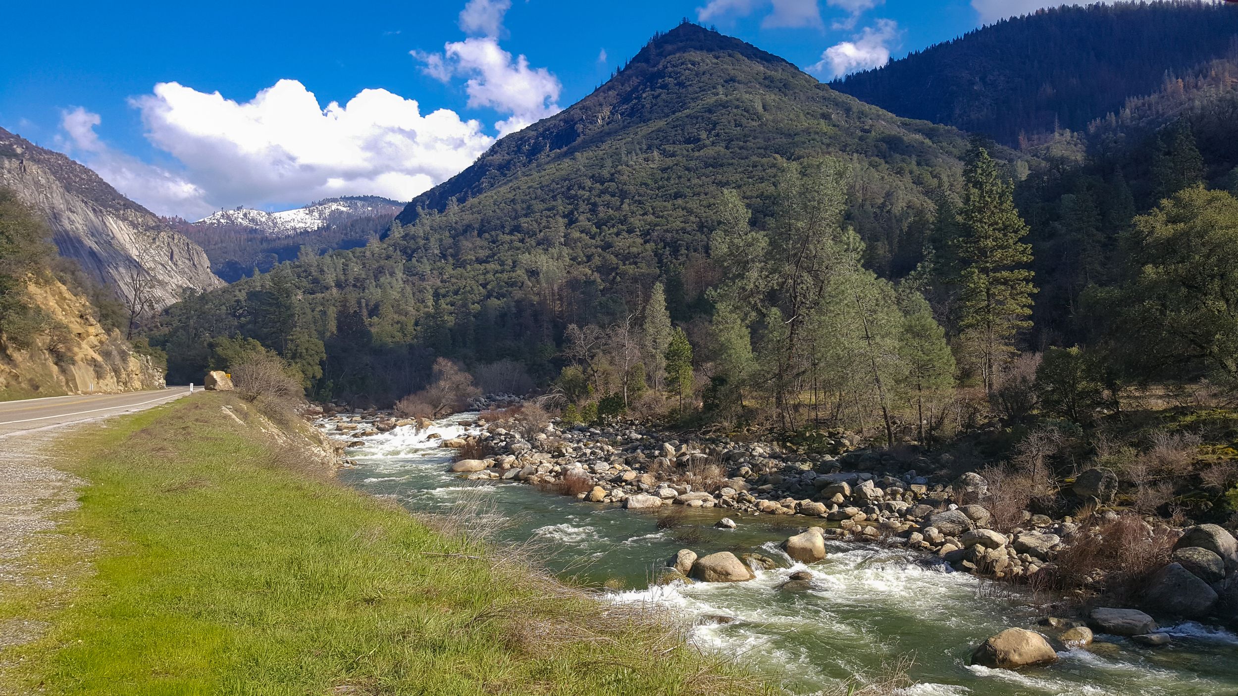 Mountain river valley with rushing waters and forested slopes under blue skies.