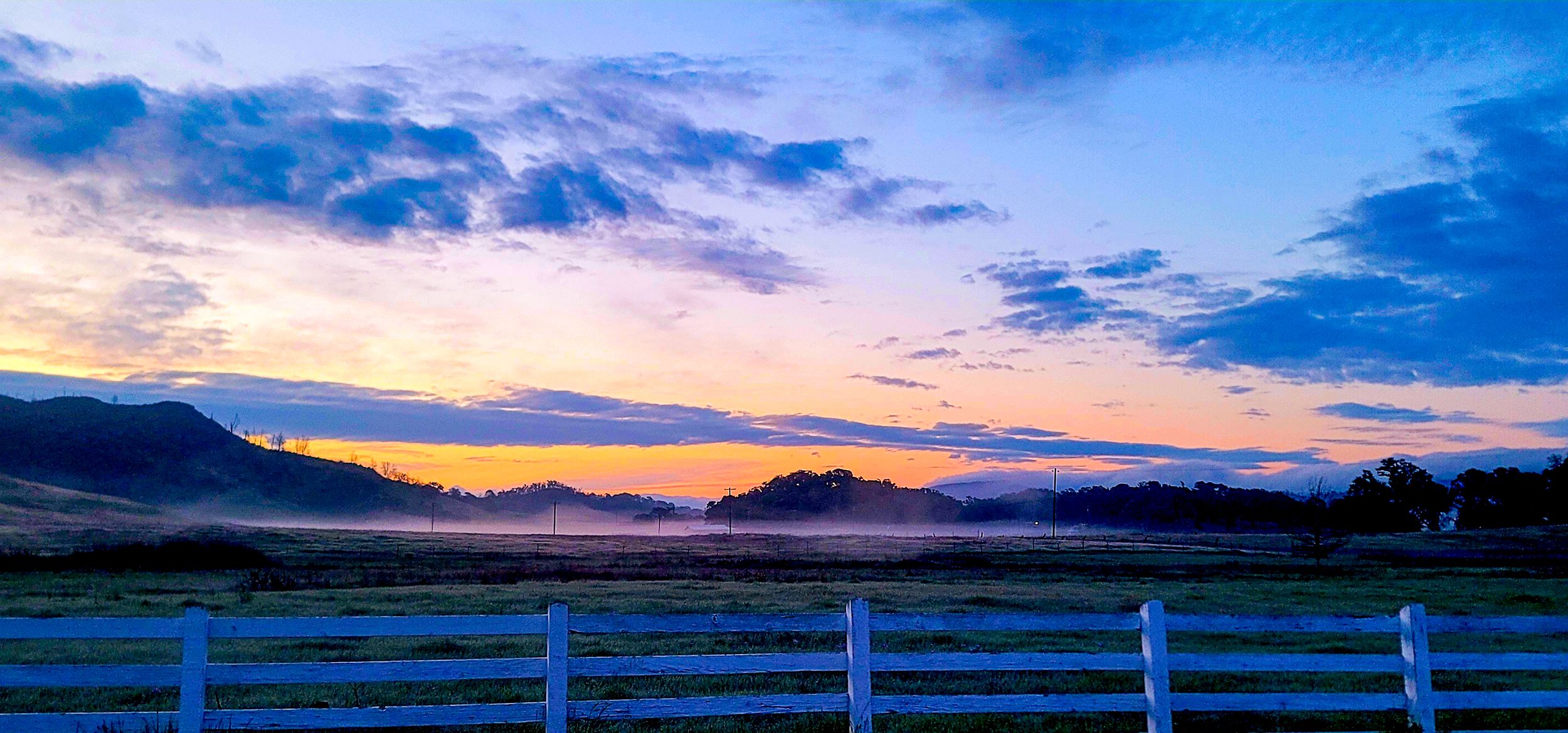 White fence along a misty field at sunset with mountains in the background.